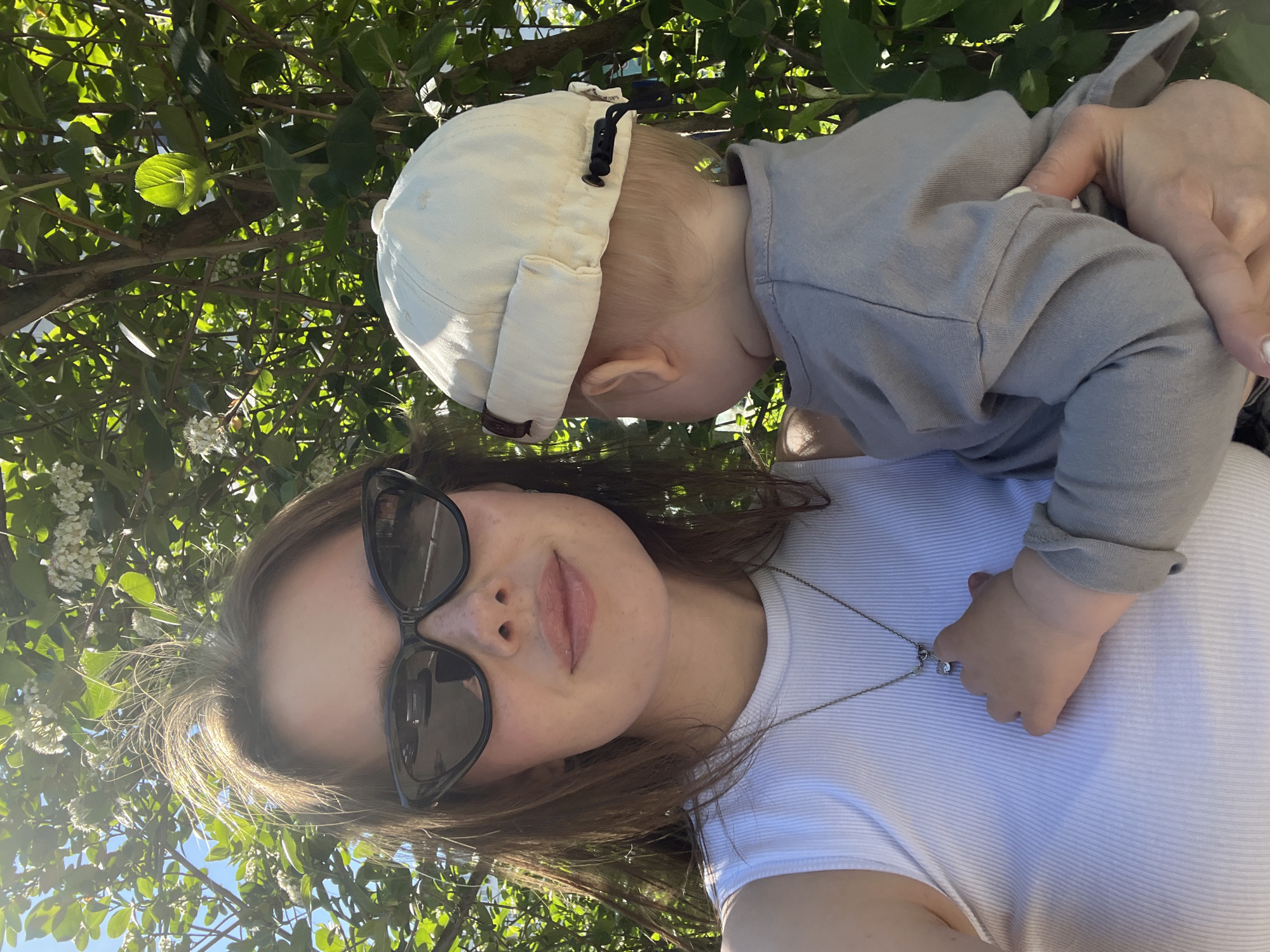 A selfie shows a woman with sunglasses and a silver necklace holding a young child wearing a white cap. The photo appears to be taken outdoors on a sunny day, with foliage visible in the background. The location is Chaikovsky, Russia.