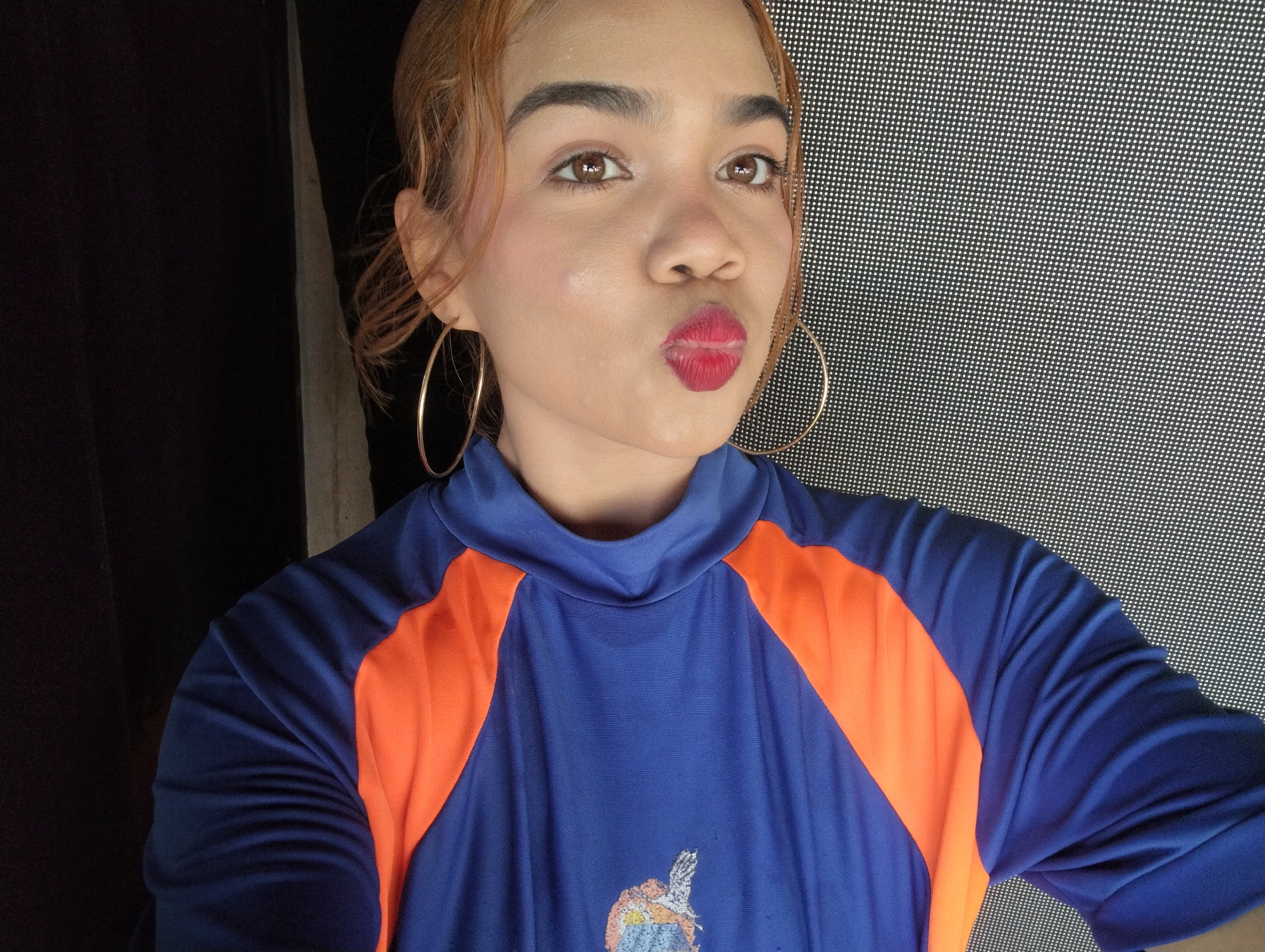 A woman is posing for a selfie. She is wearing large gold hoop earrings and a blue and orange athletic shirt. Her hair is light brown, and she is wearing red lipstick. The background is a screen with small holes. The location is unknown, and there is no information available to confirm the location of Porlamar, Venezuela.