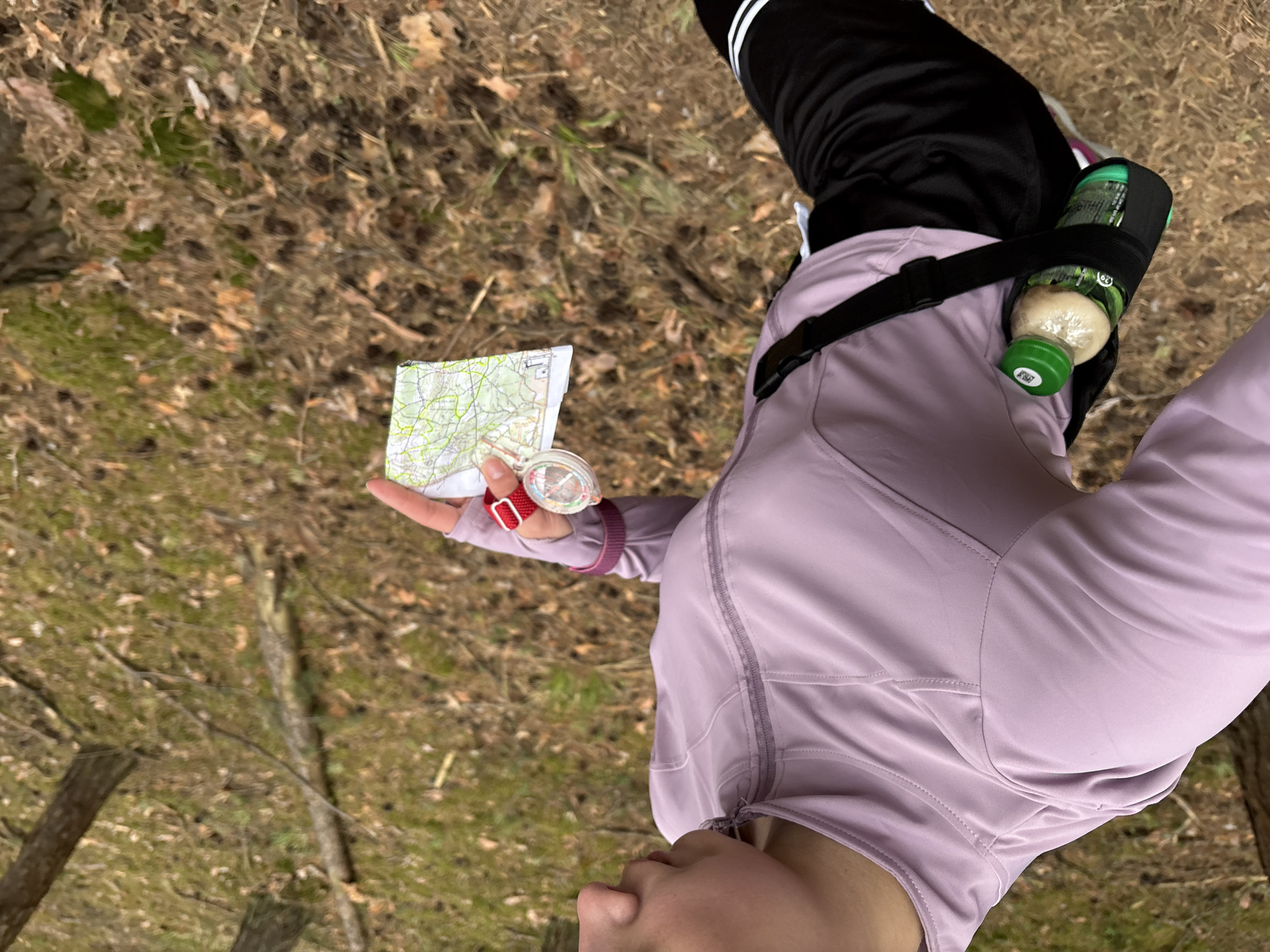 A person stands outdoors in a wooded area. The ground is covered in dry leaves, grass, and moss. The person is wearing black athletic pants with white stripes, a light purple jacket, and a black strap around the waist. They hold a map and compass in their left hand. A green-capped bottle is attached to the black strap. The location is not discernible as being within the city of Perm, Russia.