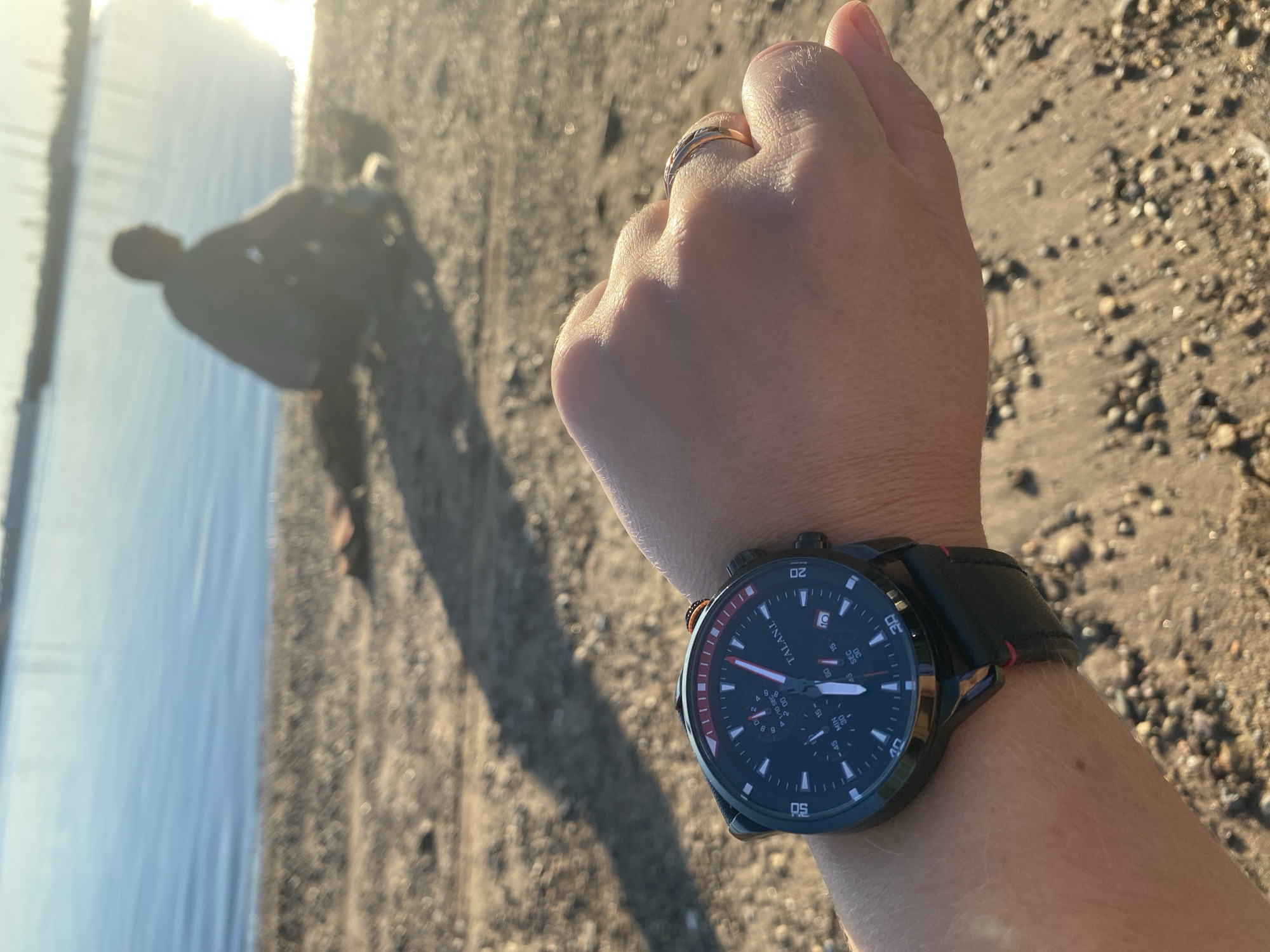A person's wrist wearing a black wristwatch with a red bezel and black leather strap is in the foreground, angled towards the viewer. The watch face displays the brand "TALANI" and has multiple sub-dials and markers. The person's hand is visible, with a ring on one finger. The background shows a sandy beach with pebbles and a body of water, possibly a river or lake, reflecting the sunlight. A lone figure is seated on the beach in the mid-ground, facing away from the camera. The scene appears to be outdoors in daylight, with a warm, golden light suggesting either early morning or late afternoon. The location is identified as Noviy, Russia.