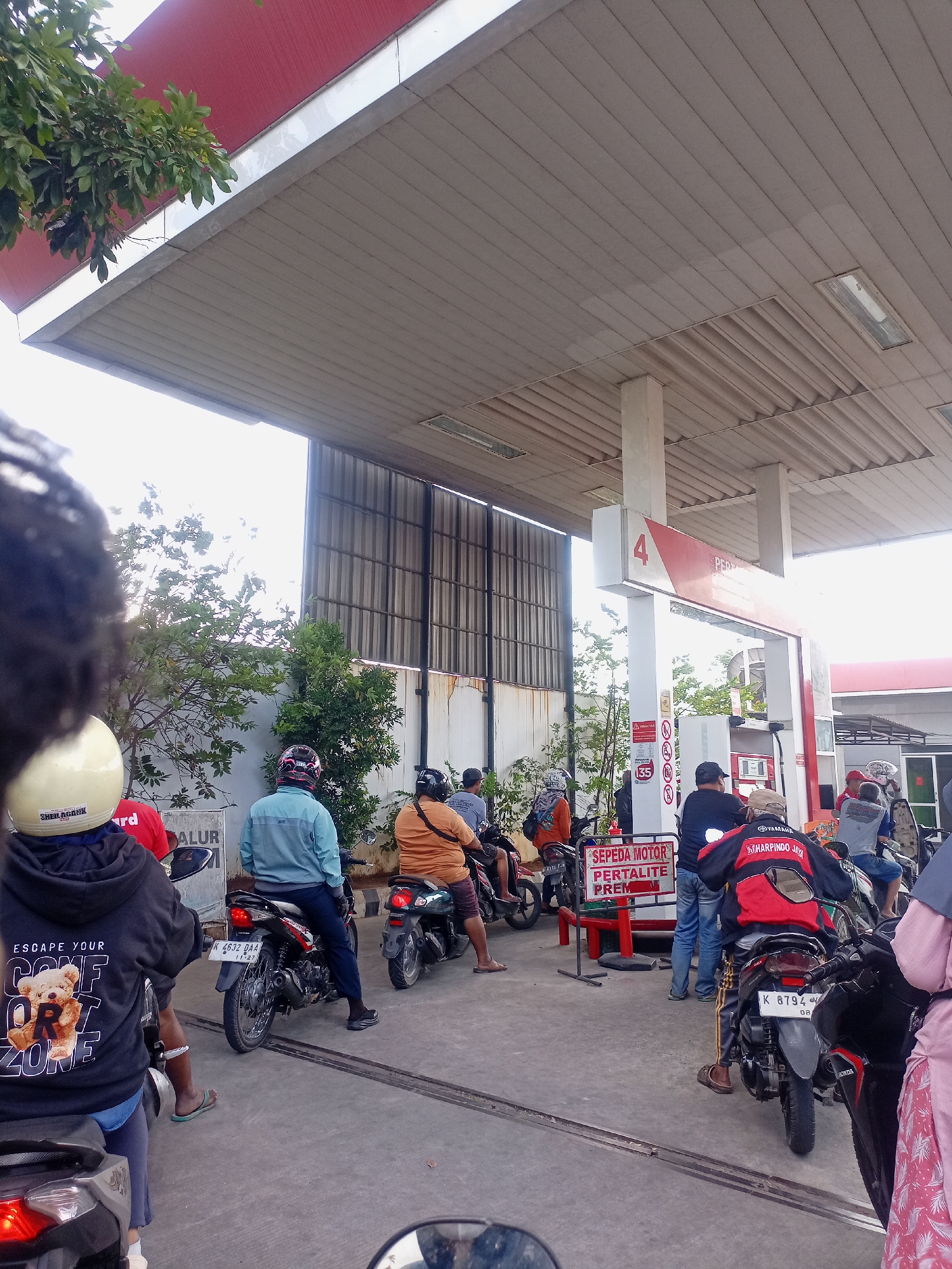 A group of people on motorcycles are queuing at a gas station in Juwana, Indonesia. The gas station has a red and white canopy, and a tall pillar with the number "4" and "Pertamina" branding. The ground is paved with concrete, and there are some trees and bushes visible. The people are wearing casual clothing, and most of them are wearing helmets. Some are looking at their phones, while others are talking to each other. The overall atmosphere is one of a typical day at a gas station.