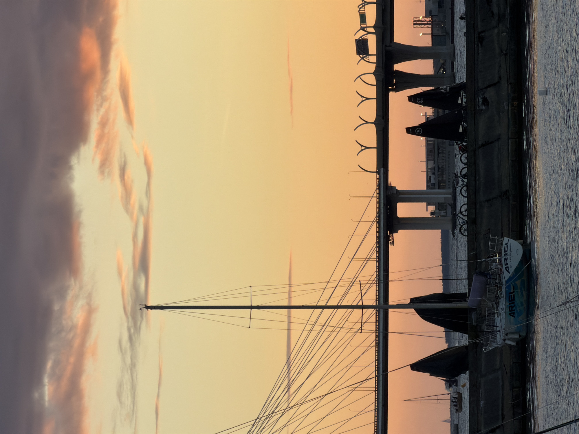 The image shows a harbor scene in St. Petersburg, Russia, during sunset. In the foreground, a white sailboat named "ARIEL" is docked. The sailboat has blue and green decals on its hull and the text "ST. PETERSBURG" is visible on the stern. Behind the sailboat, there are several smaller boats with dark sails, and a large modern bridge spans the waterway in the background. The sky is filled with dramatic orange and purple clouds. The overall atmosphere is serene and picturesque.