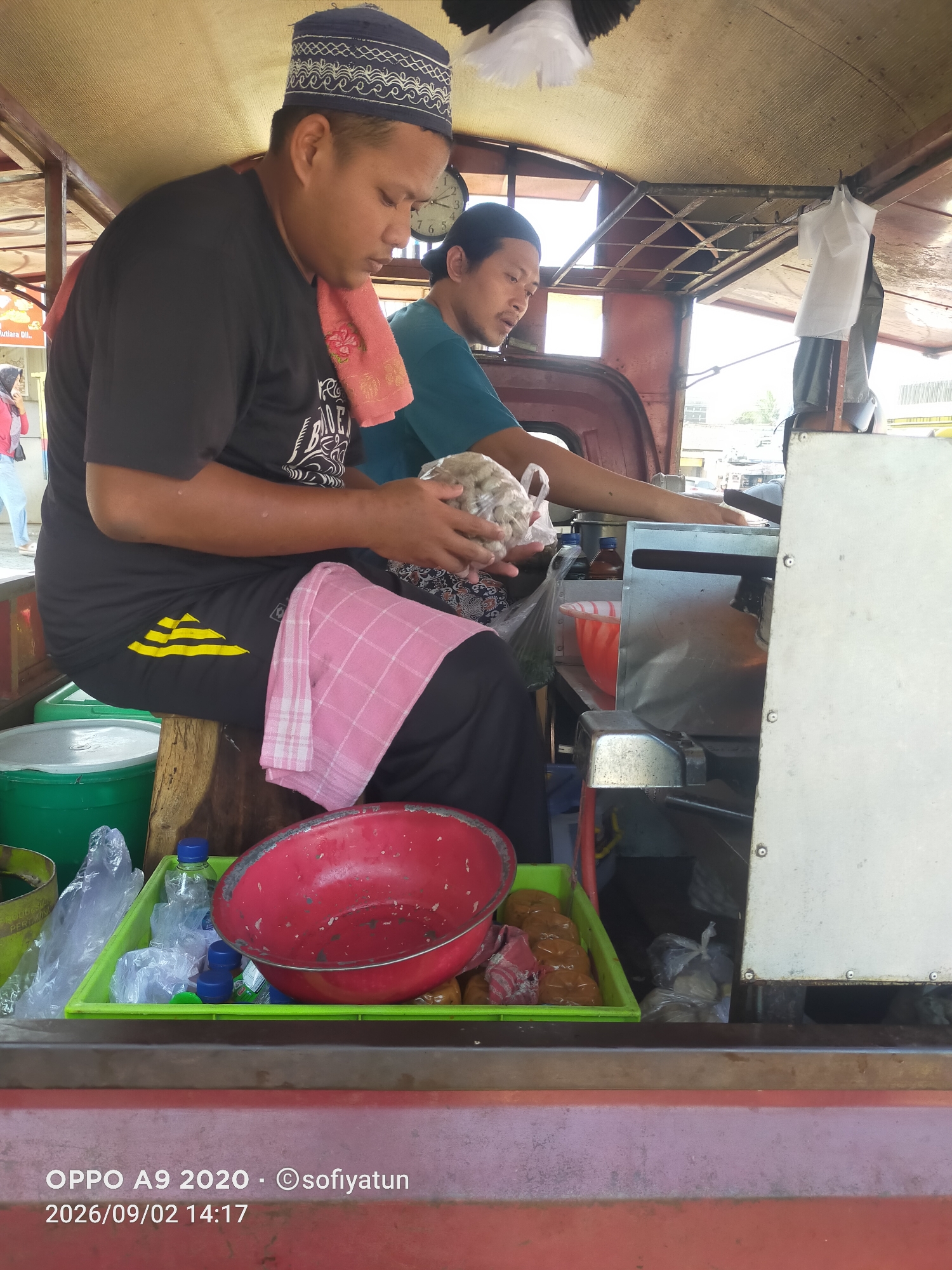 The image depicts a street food vendor stall in Tayu, Indonesia. Two men are visible, seemingly preparing food. The man in the foreground, wearing a black t-shirt and a blue peci (traditional cap), is seated and holding a plastic bag filled with what appears to be raw ingredients, possibly meatballs or fishcakes. A pink checkered towel is draped over his lap. To his right, another man in a teal shirt is also engaged in food preparation, though his actions are less clear.
The stall is equipped with various food preparation items. A large red bowl is prominent in the foreground, situated in a green plastic tray containing bottles and plastic-wrapped items. Behind the men, a stainless steel food preparation surface and what looks like a cooking station are visible. The overall environment suggests a bustling street food operation, characterized by the natural lighting and the utilitarian nature of the equipment. The presence of a clock in the background indicates the time of day. The timestamp on the image suggests the photo was taken on September 2, 2026, at 14:17.