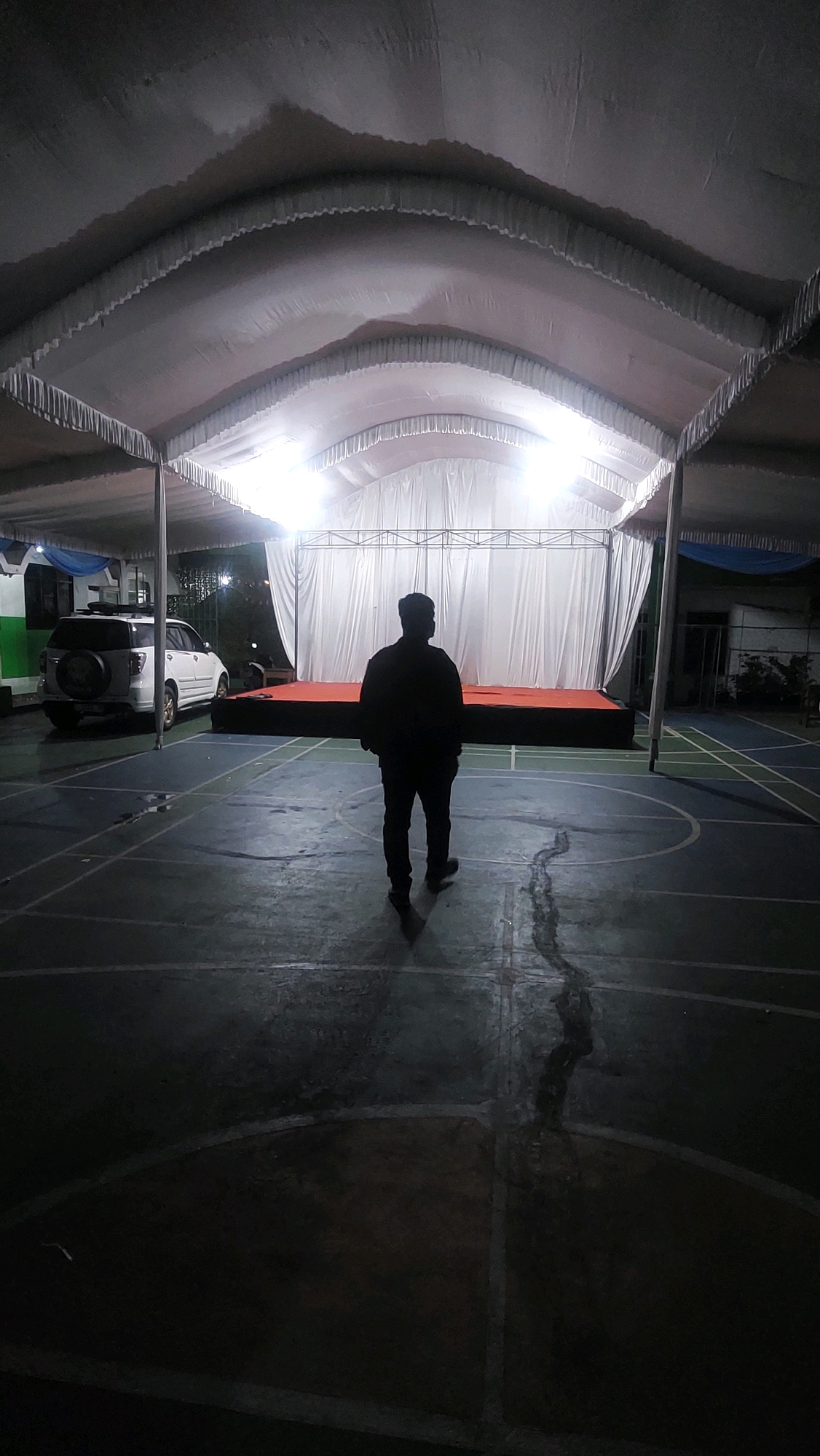 A dark, silhouetted male figure stands in the center of a basketball court, facing away from the camera towards a raised stage draped with white fabric and illuminated by bright lights. The stage is covered with a red carpet. The court has visible white lines and a crack running through it. Above, a large white tent structure provides a canopy, its edges lined with decorative ruffles. To the left of the frame, a white SUV is parked. The scene appears to be an outdoor event space at night, possibly in Cibinong, Indonesia, given the context of the city being mentioned.