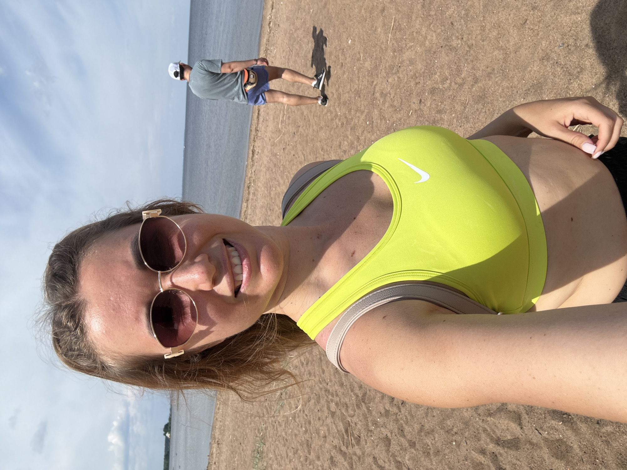 The image depicts a woman taking a selfie on a sandy beach or shoreline, with another individual in the background by a body of water under a partially cloudy sky. The location is in the city of Unknown, Unknown.
In the foreground, a female subject, appearing to be in her 20s or 30s, smiles directly at the camera. She wears large, gold-rimmed, pink-tinted oval sunglasses. Her light brown hair is pulled back, with some loose strands around her face. She is wearing a bright neon yellow Nike sports bra with a white swoosh logo on the left chest. A light beige or gray strap of a backpack or bag is visible over her right shoulder. Her left hand, with light pink-tipped fingernails, is visible at the bottom right of the frame, holding the camera or phone for the selfie. Her right arm shows a slight tan and freckles.
Behind her, to the right, a male subject walks away from the camera towards the water's edge. He wears a grey-green t-shirt, blue athletic shorts, a white baseball cap worn backward, and black and white athletic shoes. An orange fanny pack or waist bag is secured around his waist. His shadow is cast on the sand in front of him.
The environment consists of light brown sand with minimal debris in the foreground. Beyond the individuals is a calm, dark grey-blue body of water, likely a large lake or bay, extending to the horizon. In the far distance on the left horizon, a small white building or structure is faintly visible on land. The sky is largely overcast with scattered white and grey clouds, but patches of blue sky are visible, indicating bright sunlight.