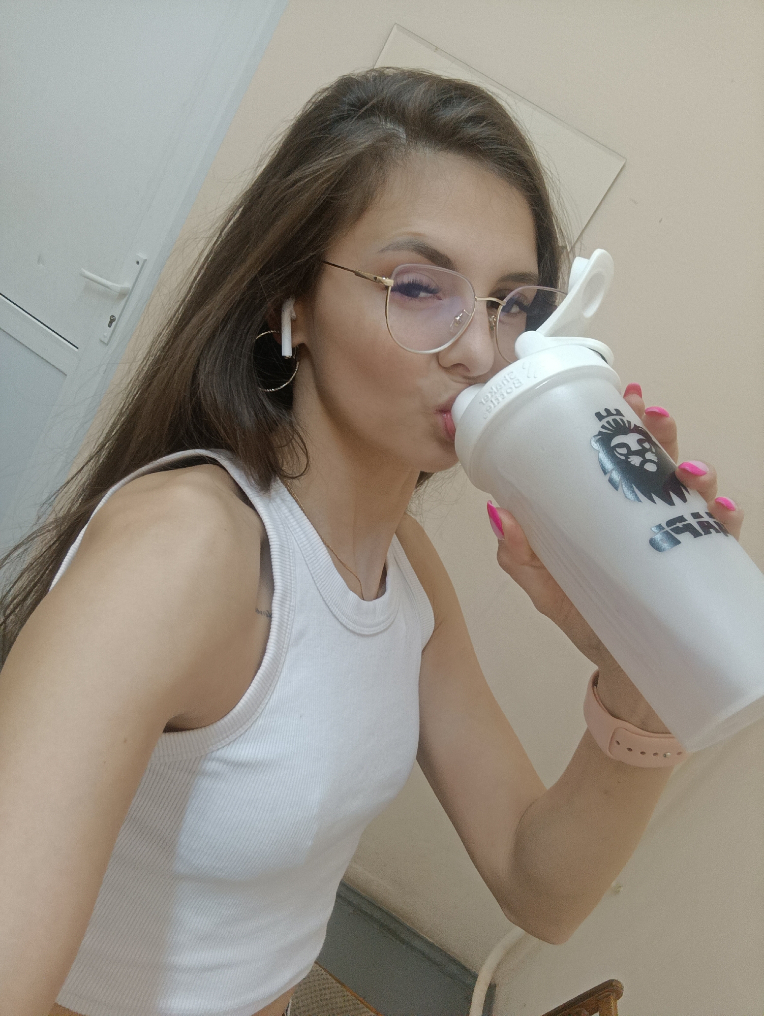 In an indoor setting, a young woman with long, dark hair and glasses is captured in a selfie. She is wearing a white ribbed tank top and has bright pink fingernails. She is holding a white shaker bottle with a black lion logo and drinking from it. The woman is also wearing hoop earrings and white wireless earbuds. The background appears to be a plain wall and a door handle, suggesting an ordinary room or apartment.