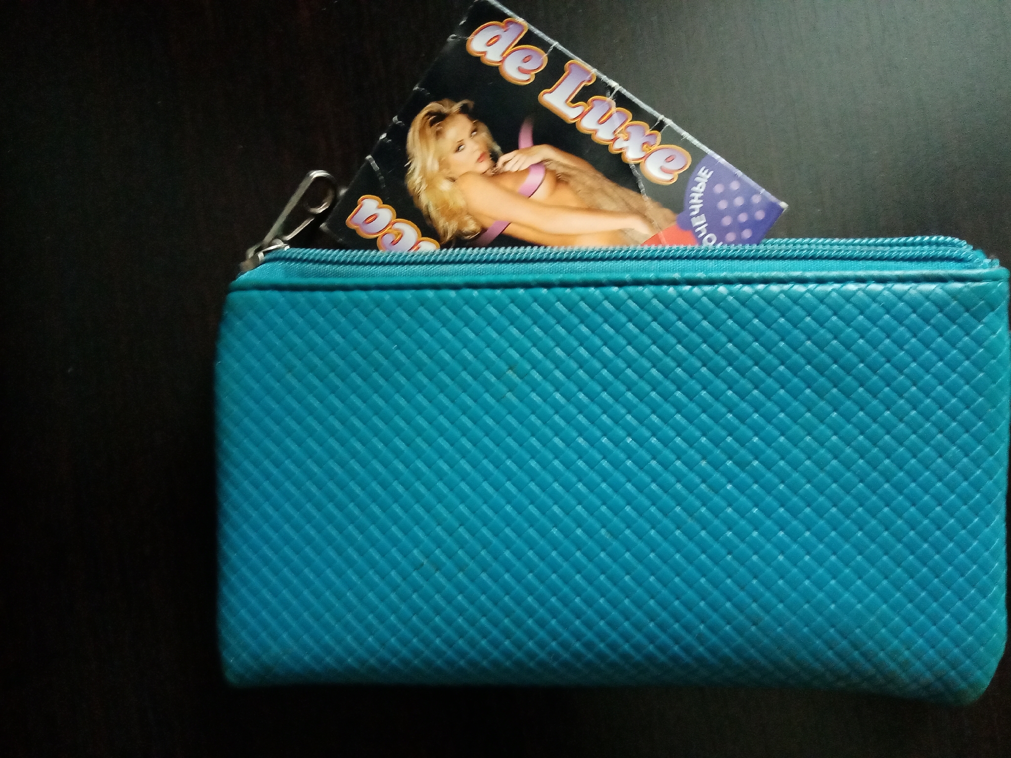 A teal or turquoise rectangular wallet, featuring a distinct woven or quilted texture, is prominently displayed on a dark, possibly black, surface. A metal zipper pull is visible on the left side of the wallet's closure. Partially protruding from the wallet is a card. The card depicts a blonde woman, likely a model, in a reclining pose, wearing a purple bikini top and gazing suggestively towards the viewer. Bold, stylized text reading "de Luxe" is visible on the card, along with partial Cyrillic text "ОЧНЫЕ," which is part of a longer word. The scene is a still life, with no active event taking place, presented under what appears to be indoor lighting. The presence of Cyrillic text aligns with the context of the image being from Elektrostal, Russia.