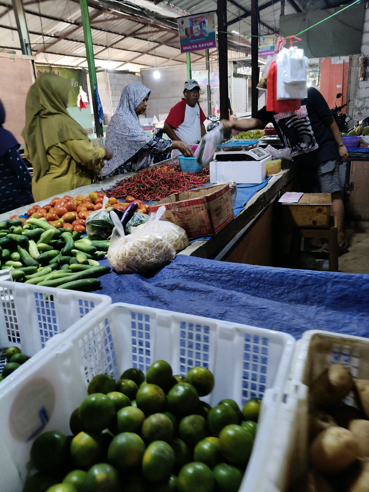 In Setiamekar, Indonesia, a bustling market scene unfolds. The image captures a vendor, partially obscured by plastic bags, handing produce to a customer at a stall. The stall is laden with fresh vegetables, including piles of red chili peppers, tomatoes, and cucumbers, alongside bags of bean sprouts and eggplants. In the foreground, white plastic crates overflow with green oranges. A man in a red and white shirt and a baseball cap is seen on the left, also attending to the market. The backdrop reveals a typical market structure with a corrugated metal roof and visible stall partitions. The overall impression is one of a lively and vibrant marketplace.