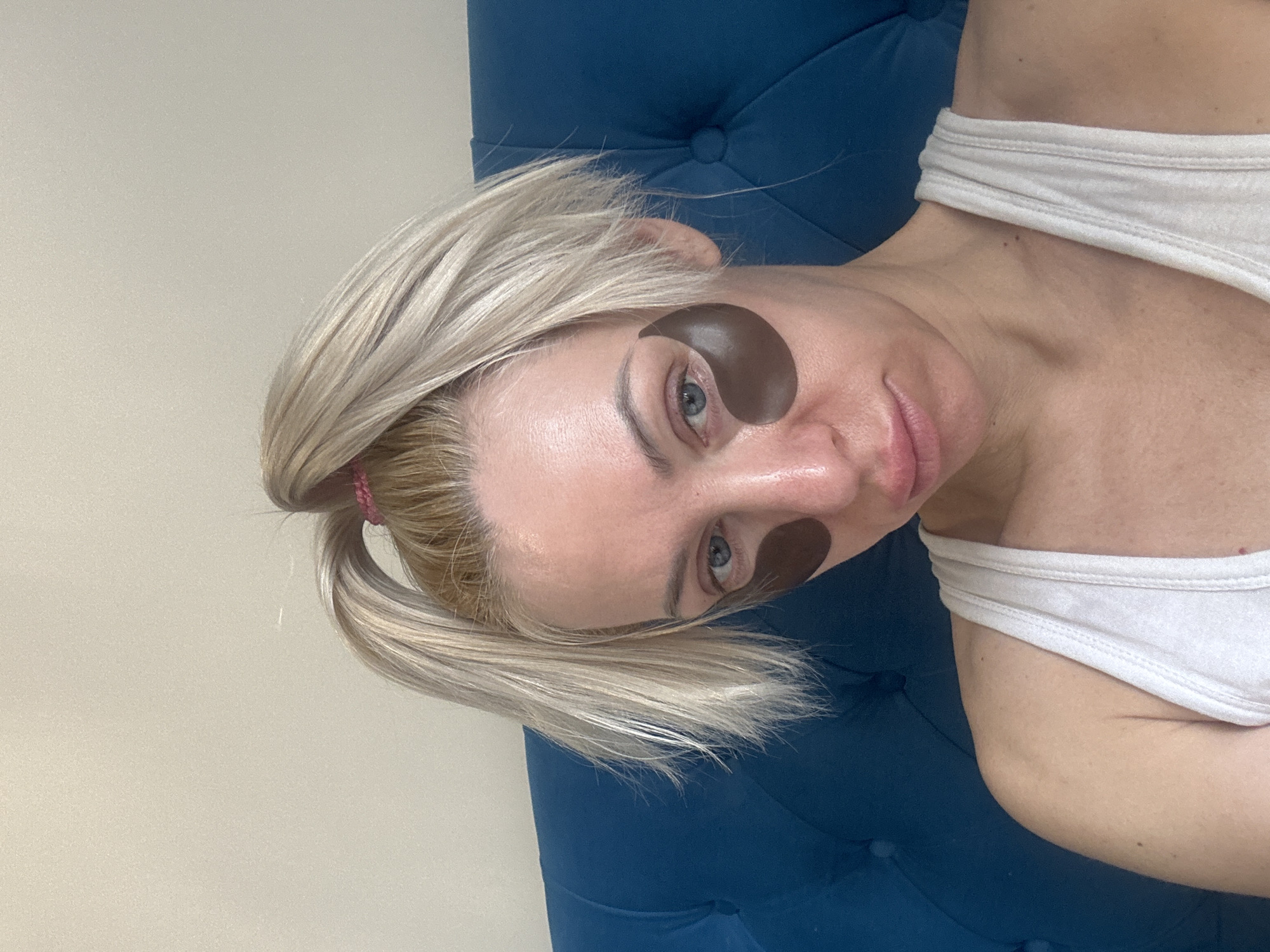A woman lies reclined on a dark blue, tufted surface. She wears a white tank top. Under-eye patches are affixed to her face. Her blond hair is pulled back in a low ponytail. The background consists of a light beige or off-white wall. The location and city are not discernible from the image.