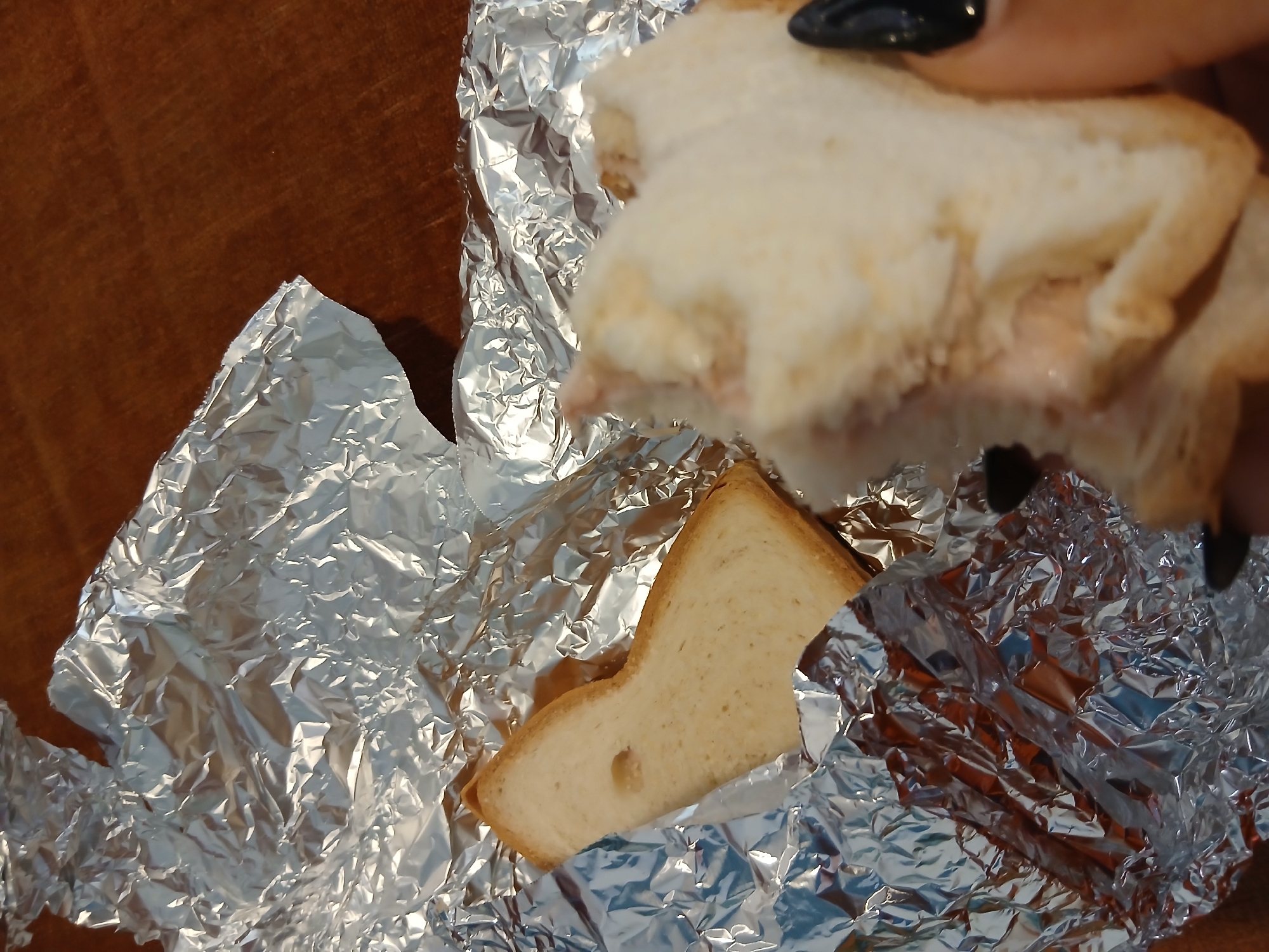 In Playa El Agua, Venezuela, a close-up shot reveals a partially eaten sandwich wrapped in crumpled aluminum foil. The sandwich, with one bite taken out, rests on a brown wooden surface. A single slice of white bread lies nearby, also wrapped in foil. The image focuses on the textures of the food and foil, with no discernible individuals present in the frame.