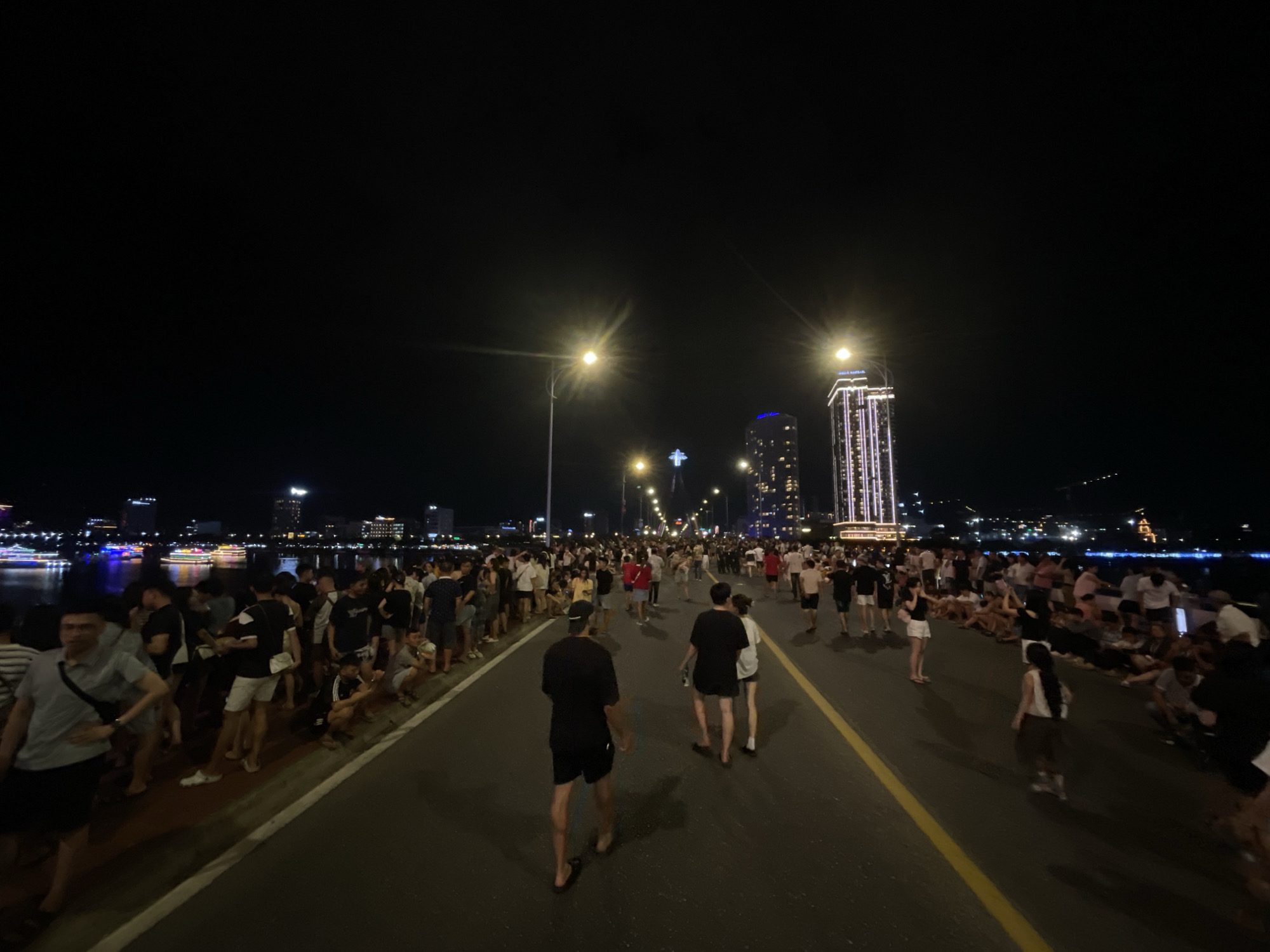 This is a night-time, wide-angle shot of a busy street in Đà Nẵng, Vietnam, likely during a public event. The scene is illuminated by streetlights and the lights of the city skyline, including a prominent tall, illuminated building that appears to be a skyscraper. A large crowd of people, dressed in casual summer attire like t-shirts and shorts, are gathered on both sides of the street and walking along it. Some people are seated on the ground or a low wall along the edge of the street, facing a body of water. The street itself has yellow lane markings. In the distance, the lights of boats and buildings are visible reflecting on the water, suggesting the scene is near a river or bay. The atmosphere appears to be festive and crowded, with many individuals observing something happening ahead or simply enjoying the ambiance.