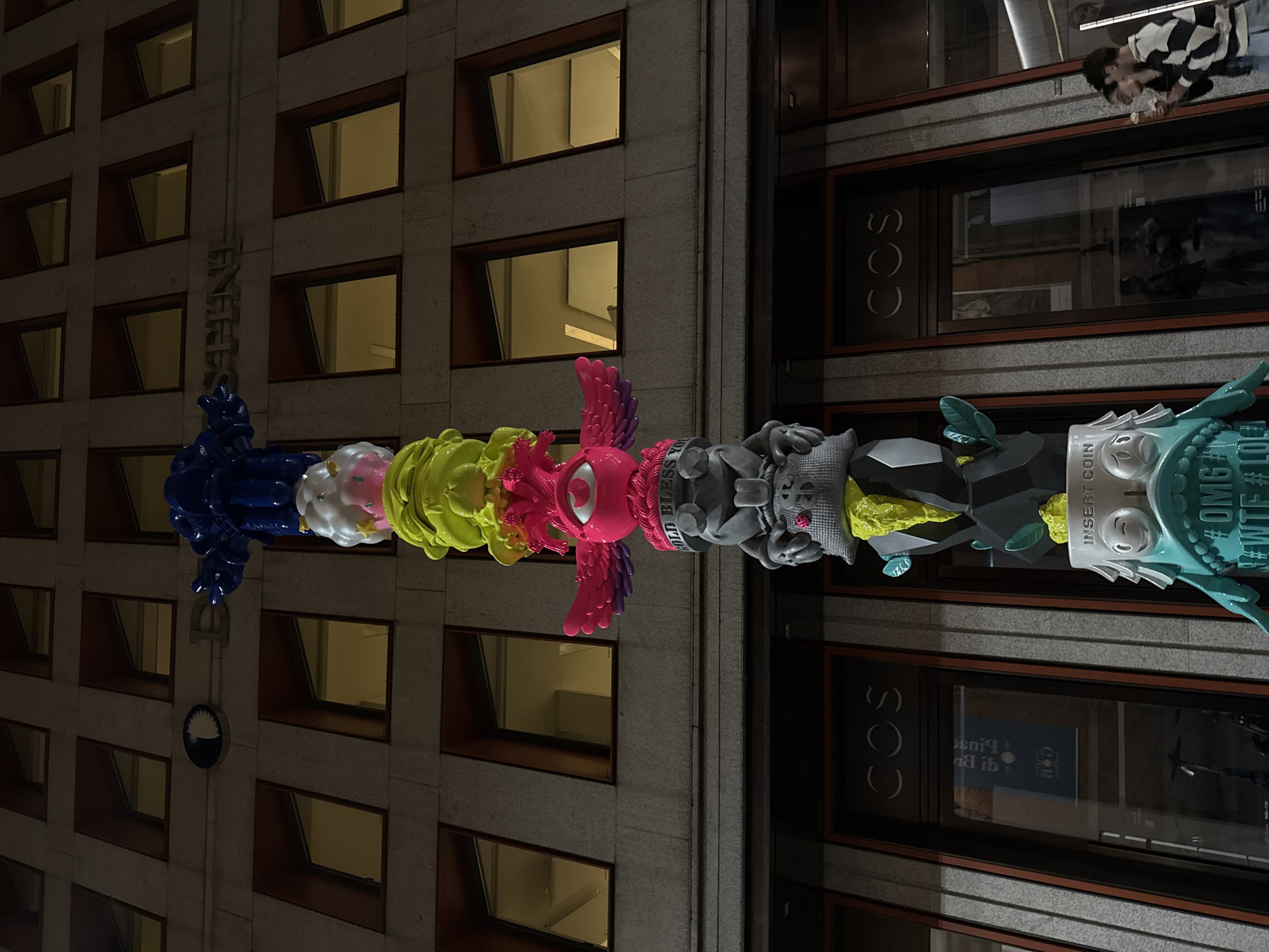 A tall, vertical sculpture composed of cartoonish, colorful figures is situated between two buildings in Milan, Italy, during nighttime. The sculpture consists of a stack of character heads, each with a unique design and color scheme. From left to right, there's a blue abstract figure, a white head with cloud-like details, a neon-green figure, a pink head with a single eye and feathery wings, a grey figure with text on its head, a grey figure with a yellow sprout, a dark grey figure with foliage, and a turquoise figure with the text "#OMG #WTF".
On the left, the sculpture is positioned against the facade of a building with numerous windows lit with warm light. On the right, the sculpture is placed against the storefront of a "COS" retail store. Through the store windows, reflections of the street and city lights are visible. Above the COS store, a person wearing a black and white striped garment is looking down.
The sky is dark, suggesting it is night. The scene appears to be well-lit by streetlights and building lights.
