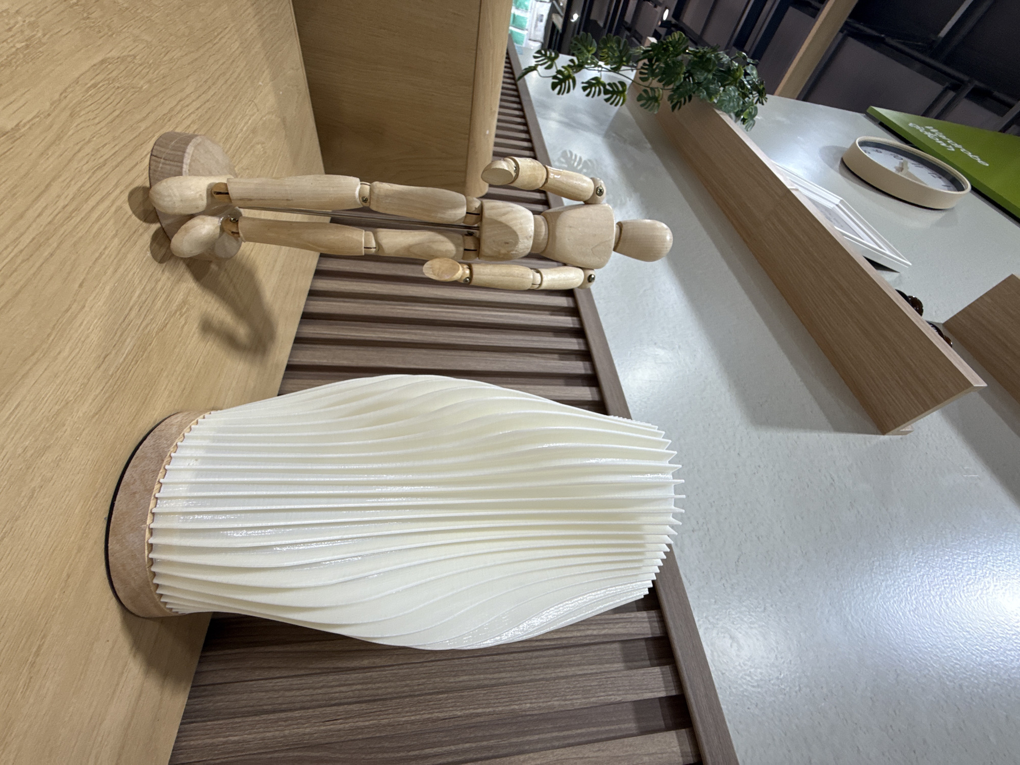 A wooden artist's mannequin stands upright on a wooden desk, its articulated limbs positioned to its sides. To the right of the mannequin, a cream-colored pleated lamp rests on the desk, its shade appearing to be made of plastic. The desk surface is light wood with visible grain. In the background, vertical wooden slats form a textured wall. Above the wall, two light-colored wooden shelves are mounted on a textured white wall. A round clock with a beige frame and black numbers is visible on the upper shelf, along with a framed picture. A potted plant with green leaves hangs from the left side of the upper shelf. The scene is brightly lit.