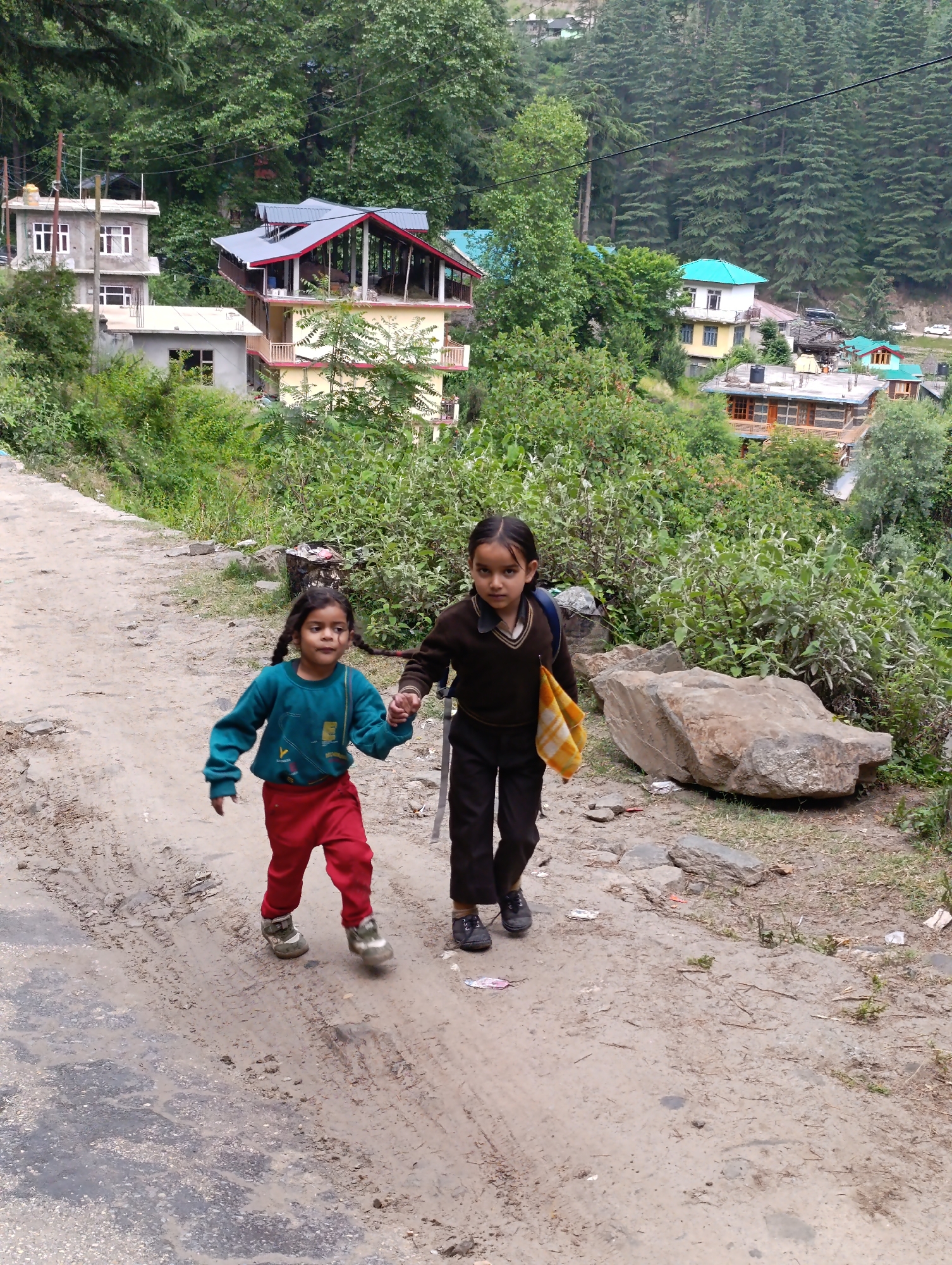 The image depicts two young girls walking hand-in-hand down a dirt and gravel path in a mountainous village, identified as Naggar, India. The scene is set on what appears to be an unpaved road or trail winding through a lush, green landscape.
The girl on the left is smaller, wearing a vibrant teal long-sleeved top and red pants. Her pigtails are visible, and she appears to be in motion, perhaps skipping or running, with a slightly blurred, lively expression. The girl on the right is taller, dressed in a brown V-neck sweater over a dark shirt, dark pants, and dark shoes, suggesting a school uniform. She carries a blue backpack and holds a yellow and orange patterned cloth in her left hand, while looking directly at the camera with a composed expression.
The path beneath their feet shows tire tracks and scattered small rocks. Large boulders are visible on the right side of the path, alongside overgrown green bushes and foliage. In the background, numerous multi-storied houses with varied architectural styles, some with flat and others with sloped roofs, are nestled amidst a dense forest of tall, green trees that blanket the surrounding mountainside. The general lighting suggests it is daytime, possibly overcast or partly cloudy, as there are no strong shadows. No discernible text is visible in the image. The scene conveys an everyday moment of childhood companionship within a picturesque, rural mountain setting.