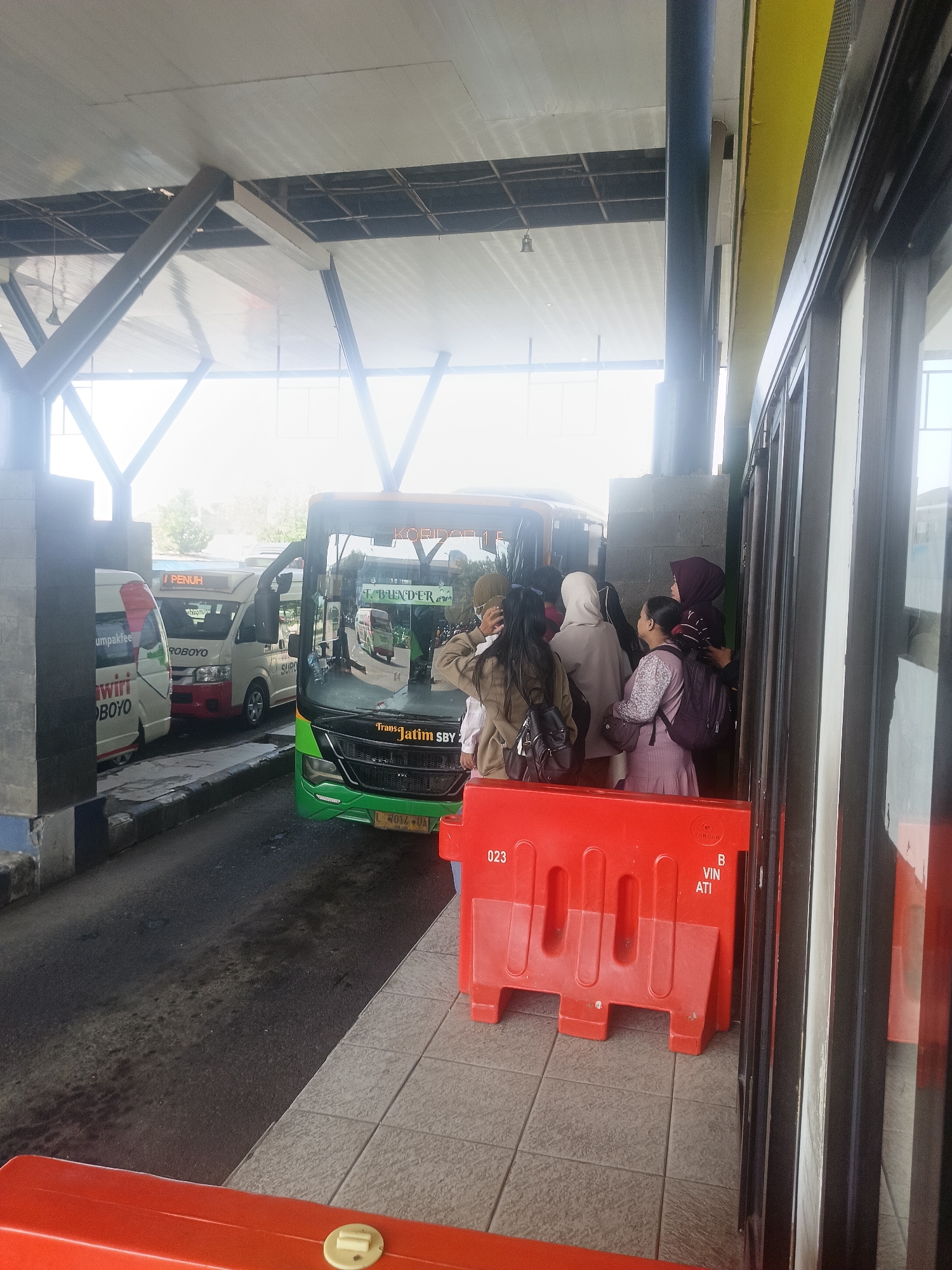 The image captures a scene at a bus station in Kedungrejo, Indonesia, possibly a terminal or waiting area. A green bus with "Trans Jatim SBY" branding is parked, with passengers boarding or disembarking. To the left, smaller vans or minibuses are also present, one with "WIRI BOYO" signage. A group of people, mostly women wearing headscarves, are gathered near the bus entrance, indicating active passenger movement. In the foreground, bright orange plastic barriers are visible, commonly used for crowd control or to delineate areas within a public space. The environment appears to be an open-air covered structure with a roof supported by metal beams, suggesting a typical Indonesian transportation hub. The lighting is bright, indicating daylight conditions.