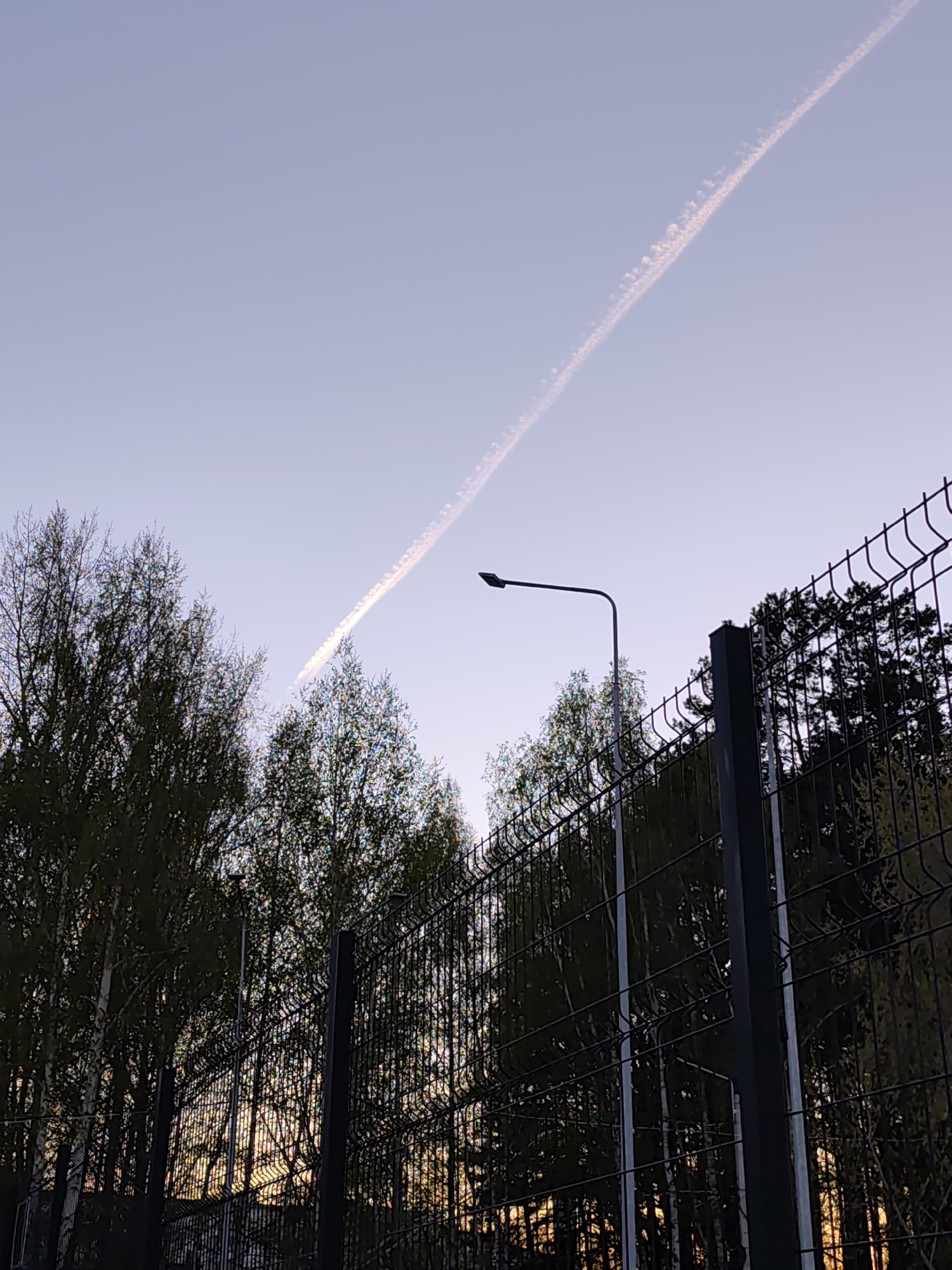 The image shows a view of a cloudy contrail in the sky above a wire fence and trees, taken in what appears to be Chaikovsky, Russia. A street light is visible in the mid-ground, positioned behind the wire fence. The trees are dark green, suggesting they are coniferous or deciduous with mature foliage. Light is visible through the trees at the bottom of the image, indicating either the setting sun or a low-lying light source. The fence is made of metal wire mesh, supported by metal poles.