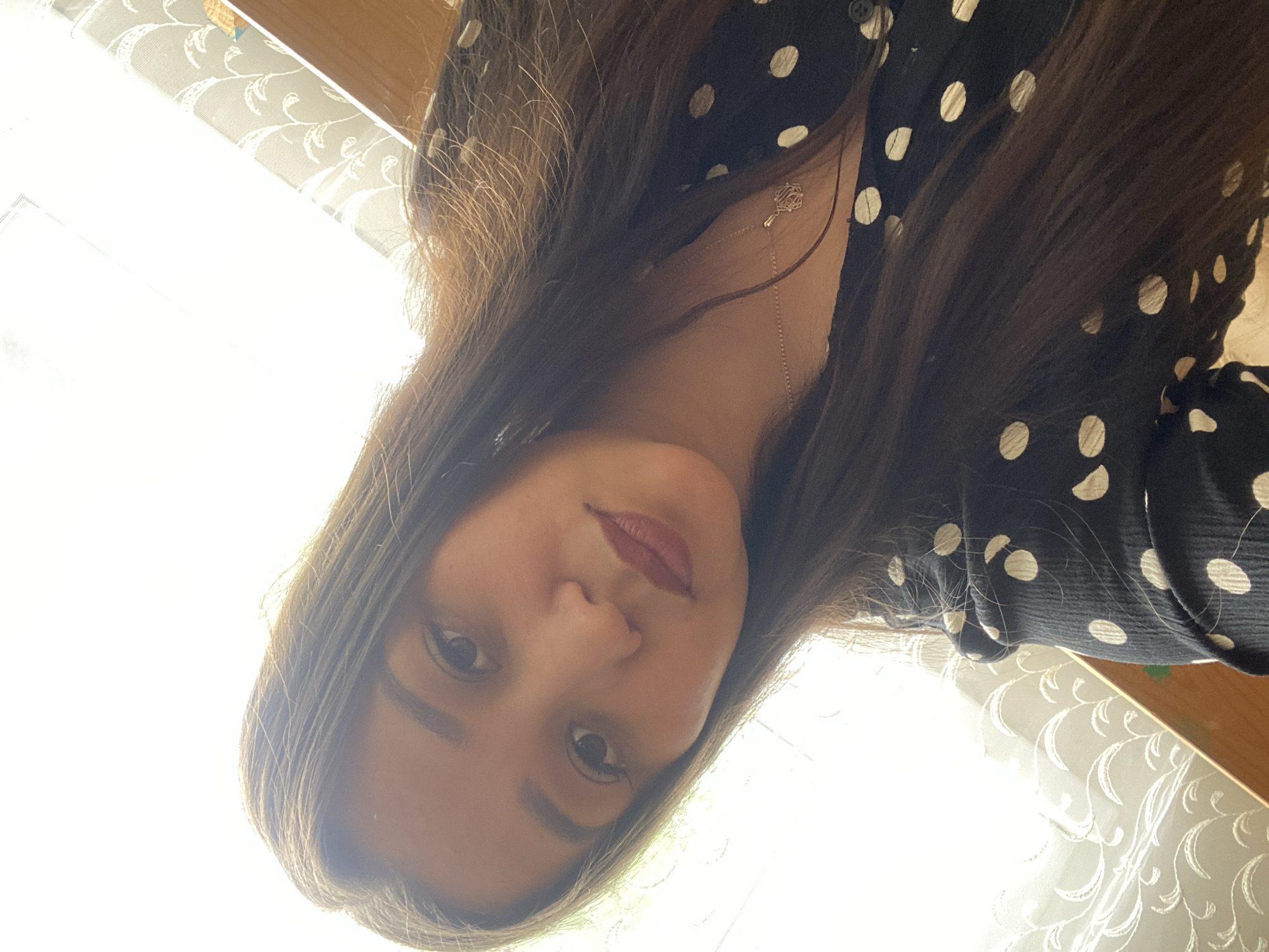 A close-up, high-angle selfie shows a young woman from the chest up, looking down and slightly to her left. She has long, dark brown hair that falls over her shoulders and chest. She is wearing a black dress with white polka dots. A thin, gold necklace with a small, intricate pendant rests on her collarbone. The background is out of focus and appears to be an indoor setting with natural light coming from a window on the left. The window has a sheer, patterned curtain. The woman's makeup includes dark eyebrows and a deep red lipstick.