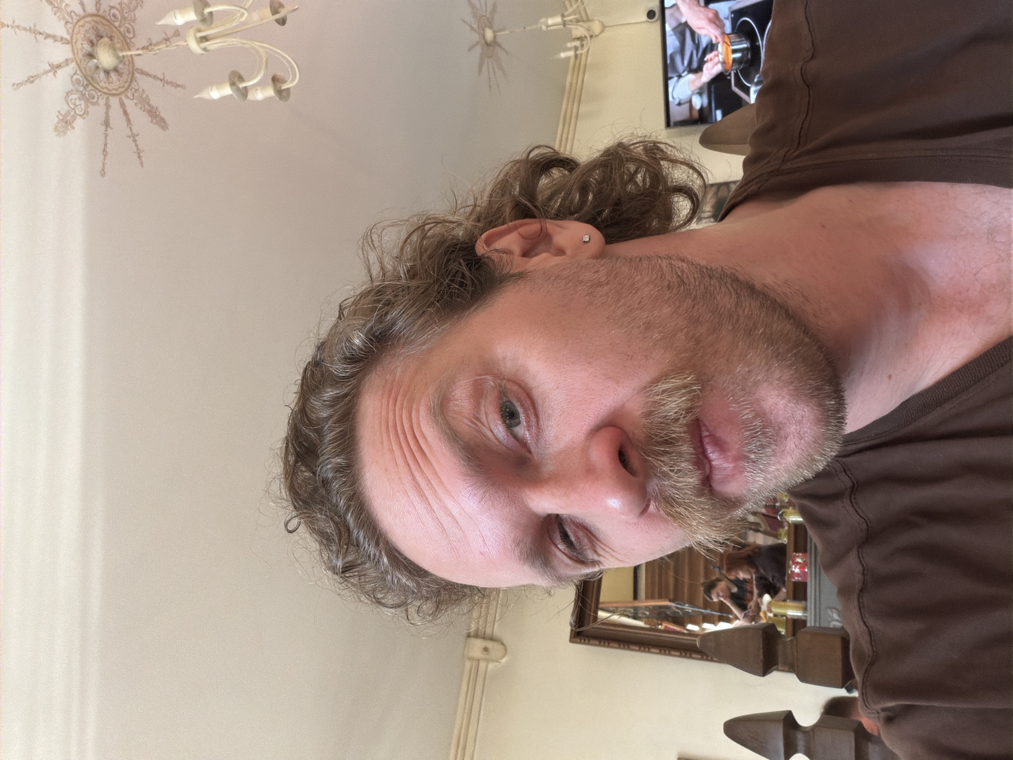A close-up, rotated shot of a man's face with fair skin, graying curly hair, and a beard. He has an earring in his left ear. The background shows a ceiling with a decorative light fixture, a TV screen showing a person working in what appears to be a kitchen, and a glimpse of a room with a mirror and other objects, suggesting an interior setting. The man is wearing a brown t-shirt. It is not possible to determine the specific location in Тэльмінскі сельскі Савет, Belarus from the given image.