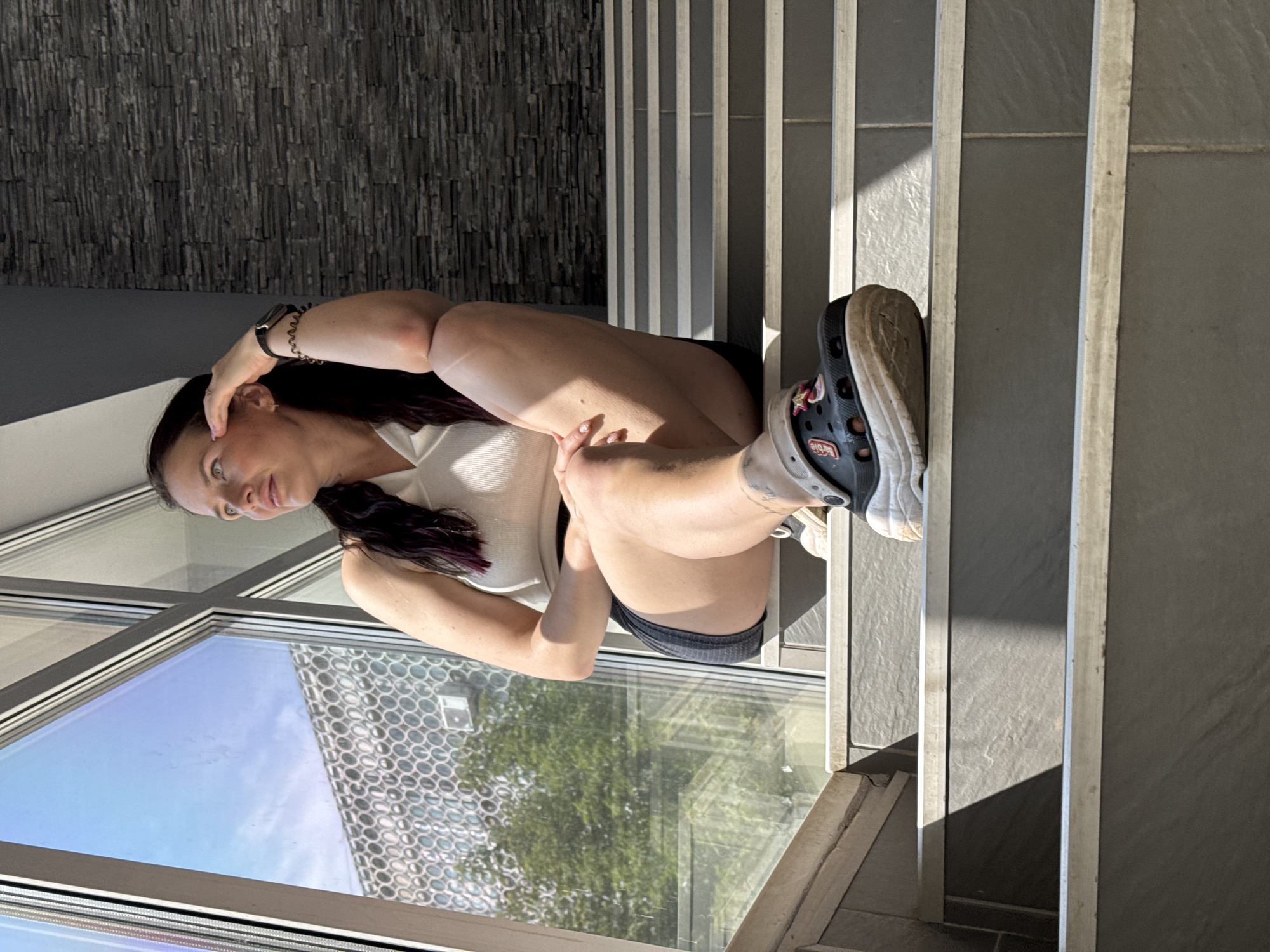 A young adult woman with dark hair, tied in braids, sits on a set of outdoor stairs. She is wearing a white tank top, dark shorts, and black crocs adorned with charms. Her left arm is propped on her knee, and her right hand is resting on her forehead. She is looking out of frame to the left, towards a large window that reflects a building with a circular patterned facade and green trees. The stairs appear to be made of stone with metal edging, and a textured dark grey stone wall is visible to her right. Sunlight casts shadows across the stairs.