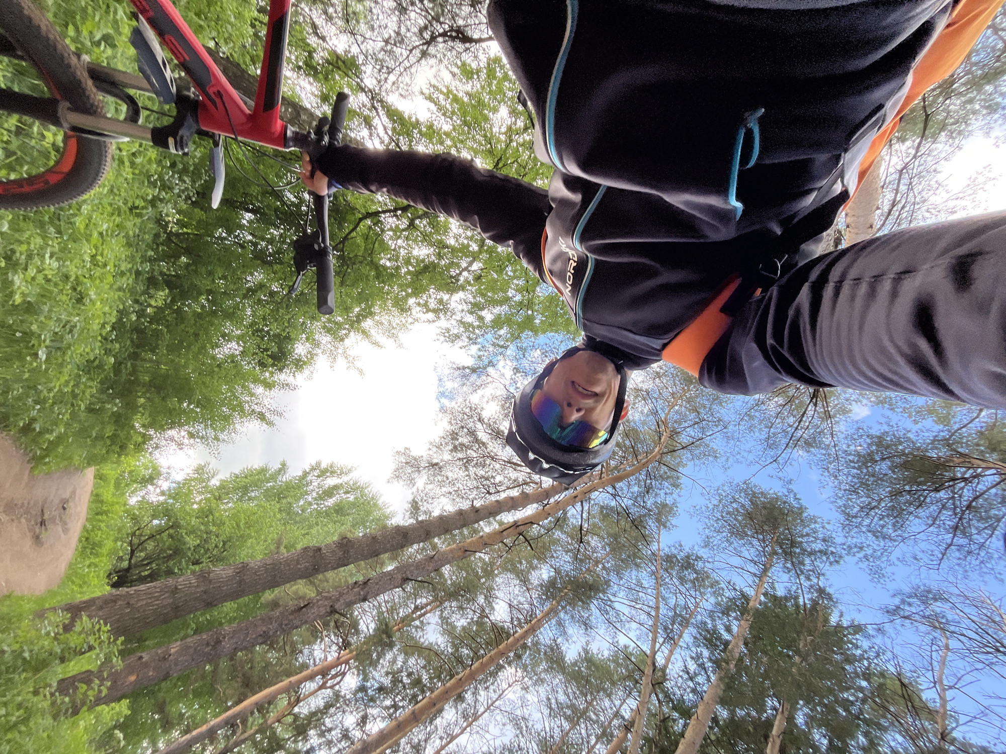 This image presents a dynamic, low-angle perspective of an individual cycling through a forest trail. The primary subject is a person, likely male, captured from below, wearing a black long-sleeved cycling jacket with teal piping and orange accents, black trousers, a black helmet, and striking rainbow-tinted reflective cycling glasses. A slight smile is visible on the person's face, suggesting contentment or enjoyment during the activity. The word "NORDIC" is partially visible on the right side of the jacket.
Parts of a red and black mountain bike are visible on the left side of the frame, including the front wheel with an orange-lined rim, a portion of the frame, and the handlebars, which the person is gripping.
The setting is a dense wooded area, typical of a forest trail in Chaikovsky, Russia. Tall trees with slender, light-colored trunks and green foliage dominate the background, reaching upwards towards a bright, partly cloudy blue sky. Lush green undergrowth and shrubs are visible closer to the ground, particularly on the left side where a dirt path or trail can also be discerned.
The photograph conveys a sense of outdoor adventure and activity on a clear, bright day, likely late morning or early afternoon. The unique perspective makes the viewer feel immersed in the scene, looking up as if from the ground or a chest-mounted camera, highlighting the scale of the trees and the movement of the cyclist.