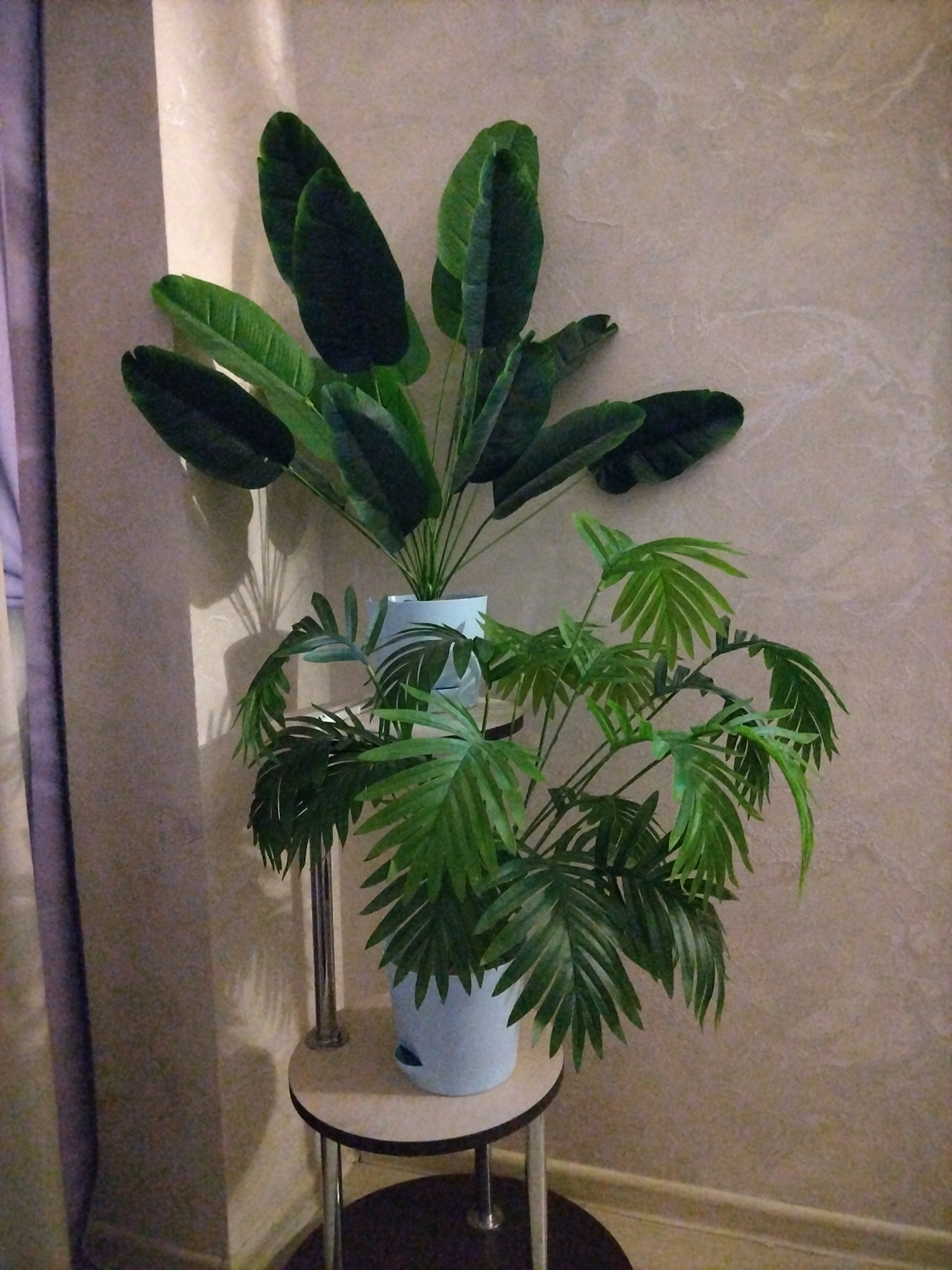 The image shows two artificial potted plants on a small circular stand, positioned near a wall with textured wallpaper and a curtain on the left. The larger plant, with broad, dark green leaves, is behind a smaller plant that has feathery, light green leaves. Both plants are in light blue pots. The stand is made of light wood and metal. The lighting creates shadows on the wall, suggesting an indoor setting. The location is identified as Rossiyskiy, Russia, though no specific urban features are visible in the image itself.