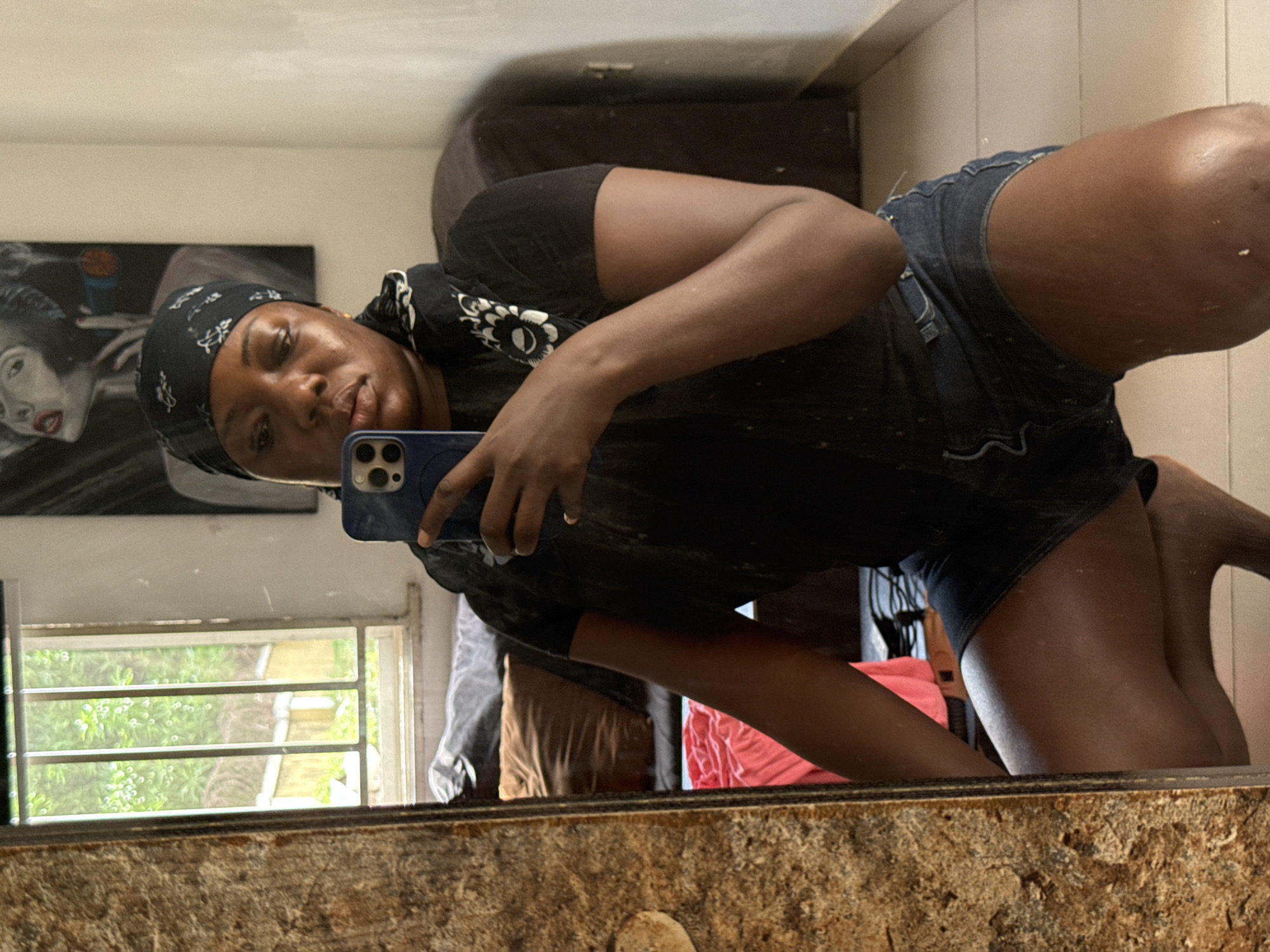 A Black woman in a black t-shirt, denim shorts, and a black headscarf is taking a selfie in a mirror. Her left hand holds a blue smartphone. In the background, there's a window with a metal grate looking out onto green trees. There's also a framed portrait of a woman on the wall. There's a dark brown piece of furniture behind the woman, and what appears to be clothing on a rack to the right of that. The location appears to be indoors. The scene does not allow for a precise confirmation of the location being in Abuja, Nigeria.