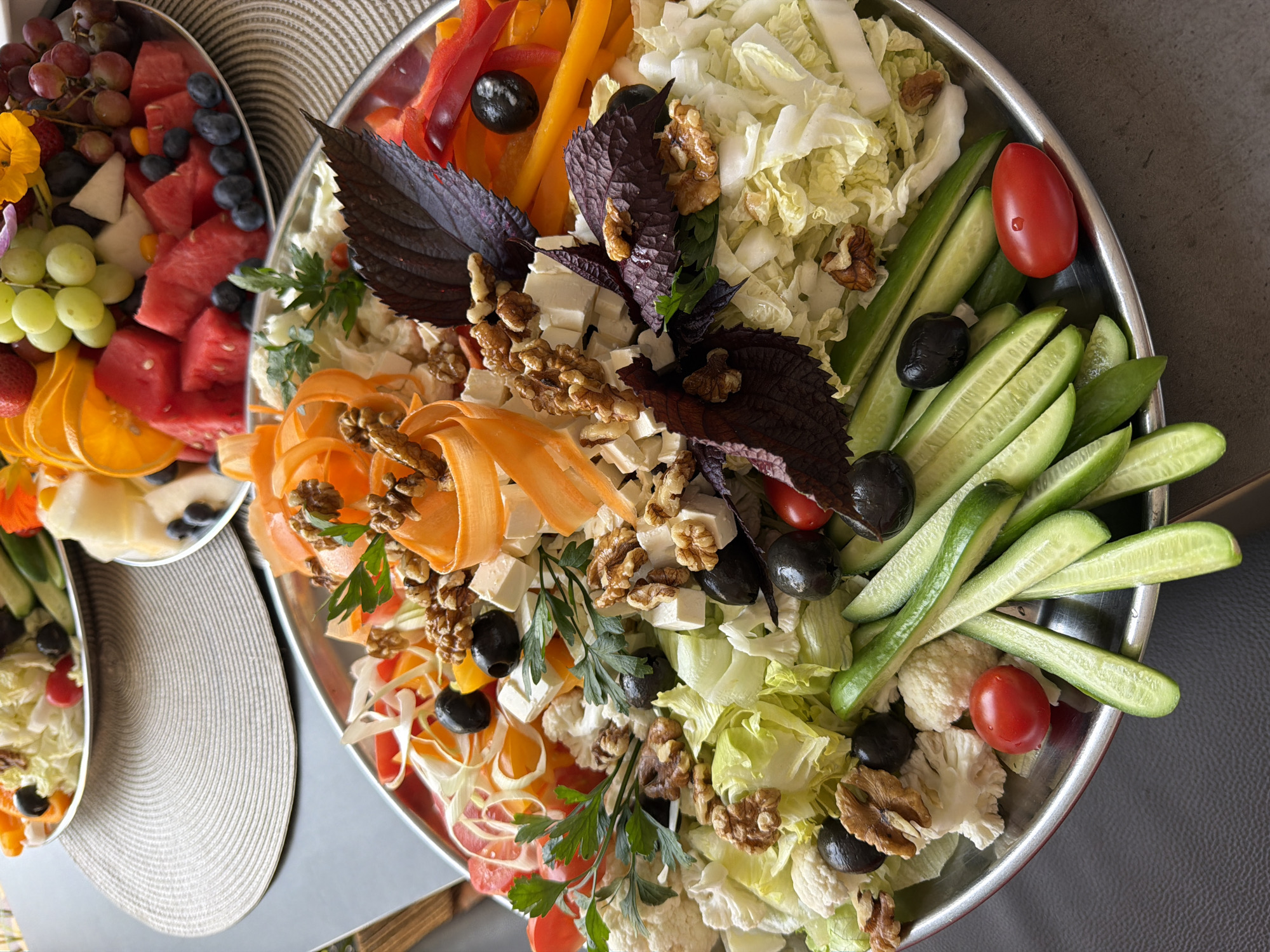 A circular silver platter overflows with a fresh salad featuring lettuce, cucumber sticks, cherry tomatoes, cauliflower florets, sliced bell peppers (red and yellow), black olives, cubes of white cheese, walnuts, and vibrant orange carrot ribbons. Dark purple basil leaves are interspersed throughout the salad. In the background, another platter contains a fruit salad with watermelon cubes, blueberries, grapes, strawberries, and orange slices. The platters are placed on a dark grey surface, with a textured grey placemat partially visible under the foreground platter. The scene is lit from above, casting soft shadows. The location is not determinable from the image.