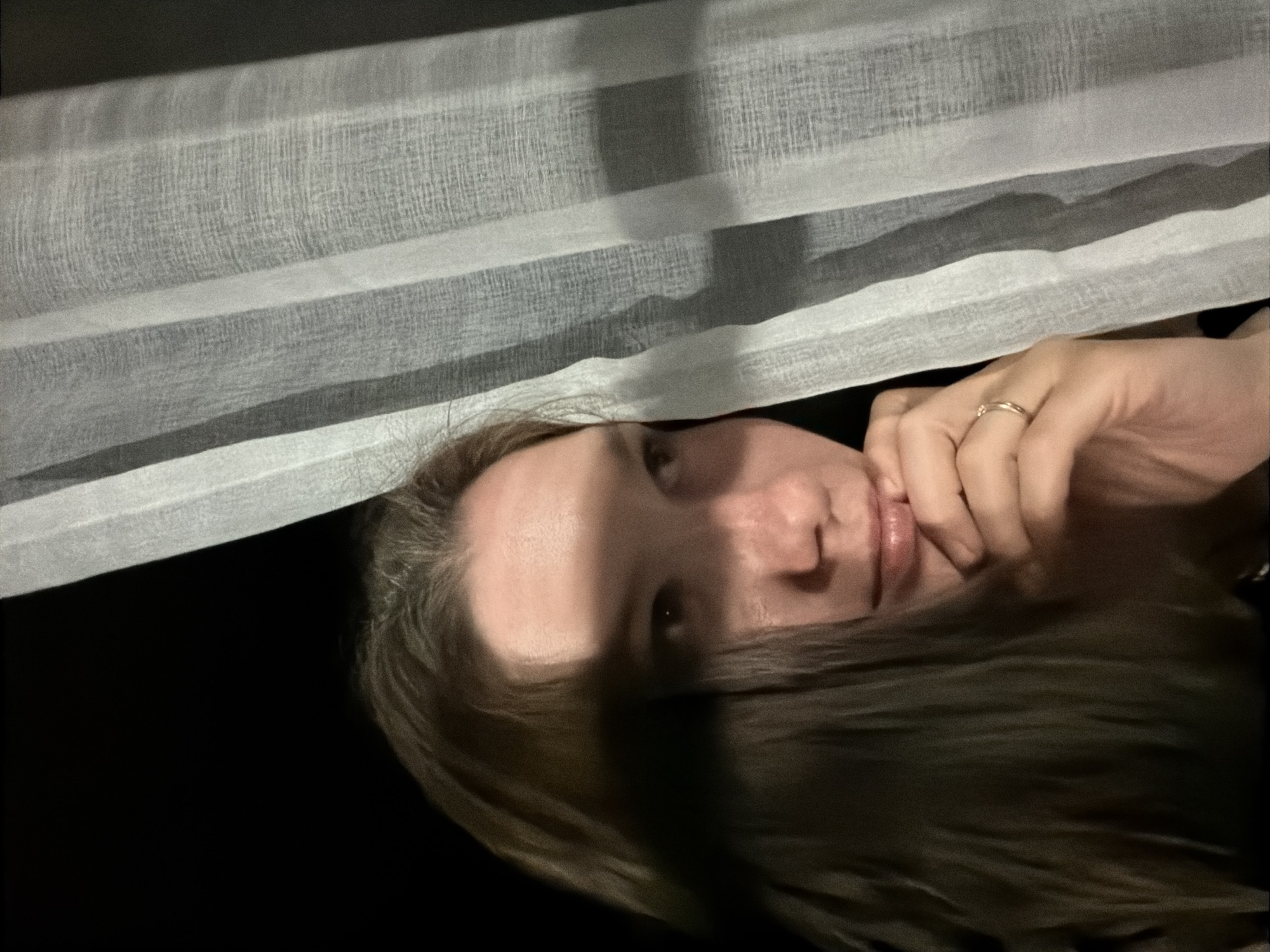 A medium shot depicts a fair-skinned person with dark brown hair. The person is positioned behind a white, horizontal curtain or blind. Light is partially blocked, casting shadows on the person's face. The individual's hand, adorned with a gold ring, is raised towards their face. The background is a dark, undifferentiated space. The image orientation is rotated. It is unlikely that specific location information such as Staraya, Russia, can be determined from the visual data provided.