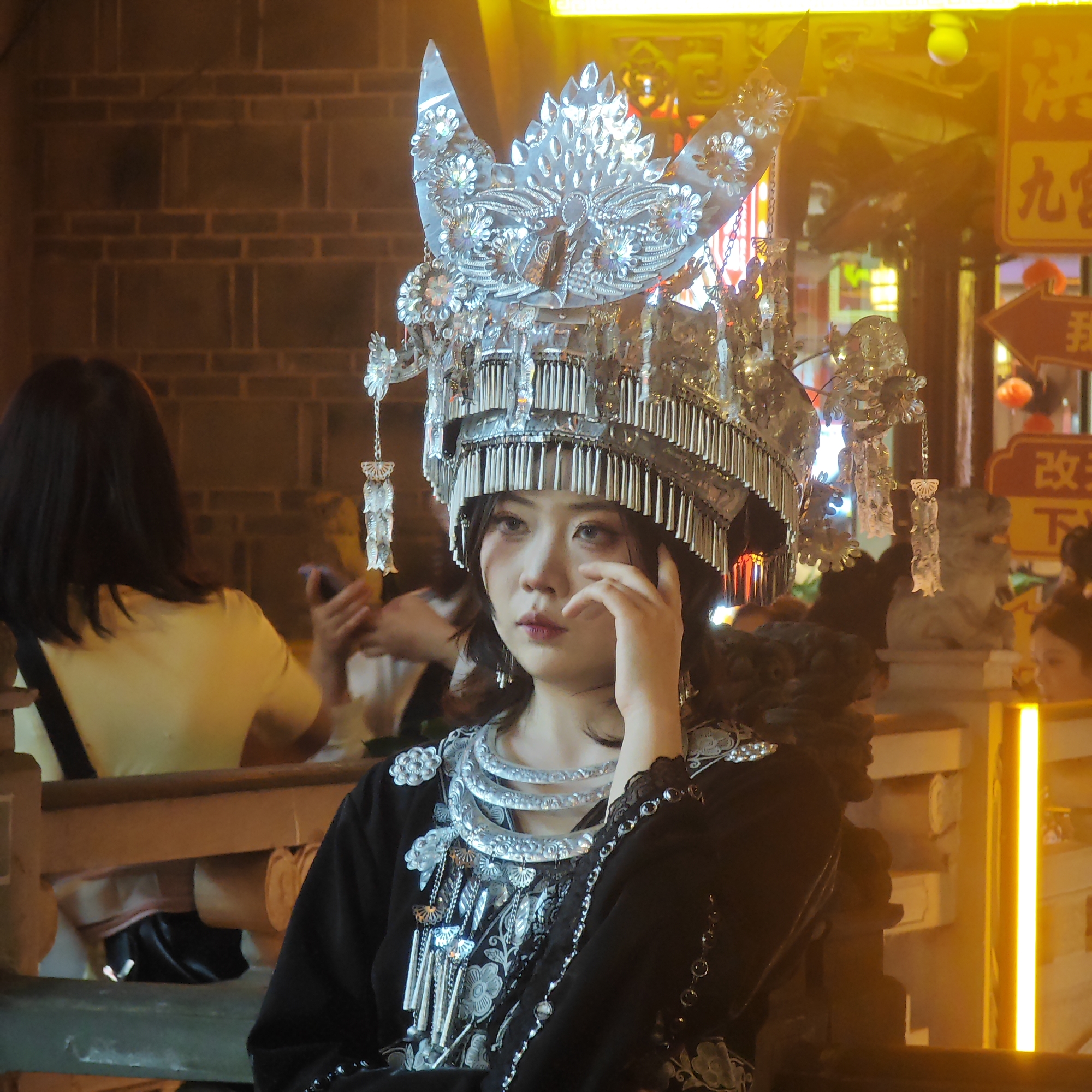 The image shows a young Asian woman wearing an elaborate, traditional silver headdress and ornate necklace. She is positioned in an urban environment, likely a commercial area, given the background signage. To her left, a woman with black hair is wearing a yellow shirt. Bright yellow lighting is visible in the background, along with brickwork on a wall. A person is visible to the right of the woman in the headdress as well as stone lion statues in the background. The area seems crowded with people. Based on the signage, it is likely that the image was taken in the Yuzhong District of China. The woman in the headdress appears to be posing, with her hand near her face.