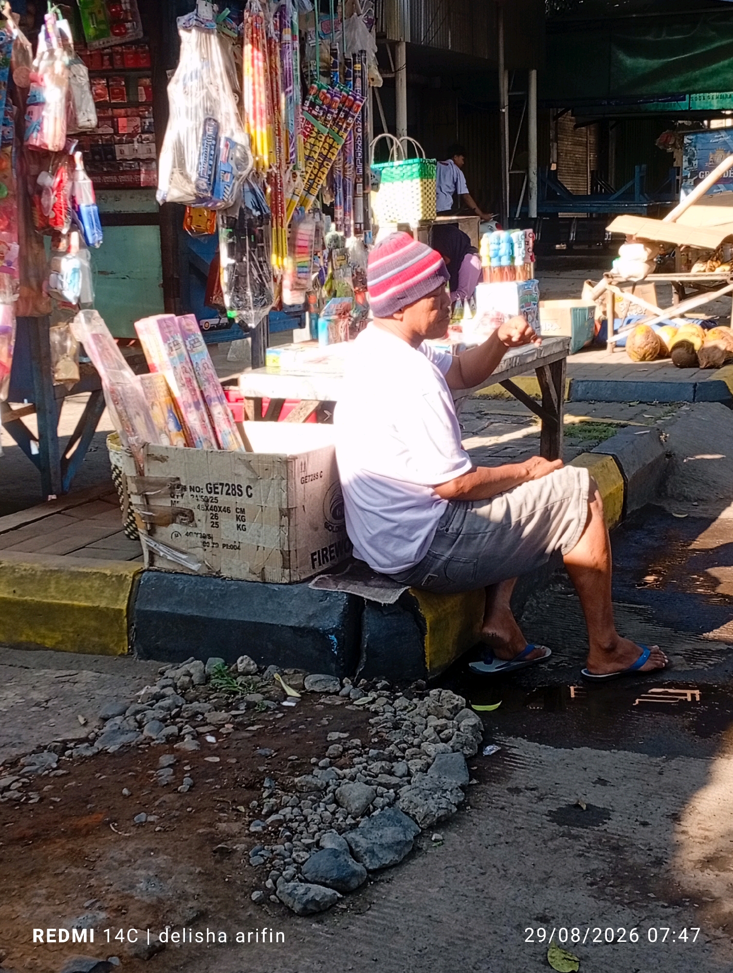 The image shows a man sitting on a curb in front of a market stall in Wonorejo, Indonesia. He is wearing a pink and white striped beanie, a light-colored t-shirt, and grey shorts. He is looking to his right and holding his right hand up. In front of him, on the ground, is a cardboard box filled with fireworks. The market stall behind him is stocked with various goods, including packaged items and what appear to be more fireworks. In the background, other market activities are visible, including people and more stalls. The ground is paved and wet in some areas, suggesting recent rain. The overall environment appears to be an outdoor market setting on a sunny day.