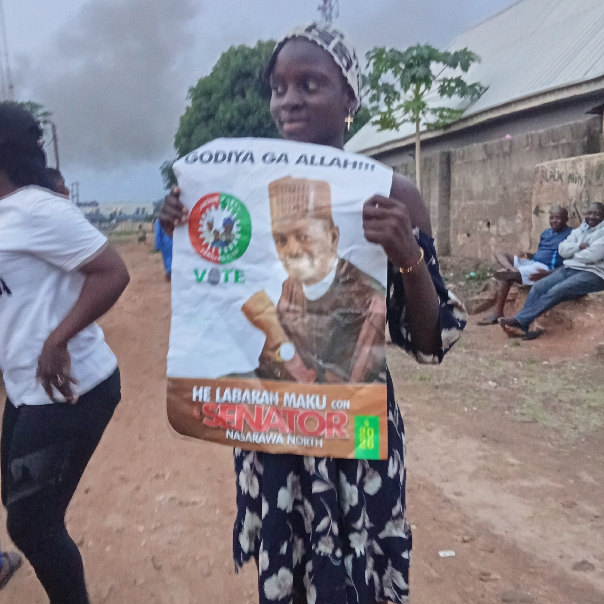 In Akwanga, Nigeria, a woman wearing a headscarf and a dress with a floral pattern holds up a campaign poster. The poster features an image of a man in traditional attire, identified as "HE LABARAN MAKU CON SENATOR NASARAWA NORTH." The poster also includes the text "GODIYA GA ALLAH!!!" and a circular emblem with the word "VOTE." Other individuals are visible in the background, some sitting on a concrete structure. The environment appears to be an outdoor, dusty area with some vegetation and buildings.