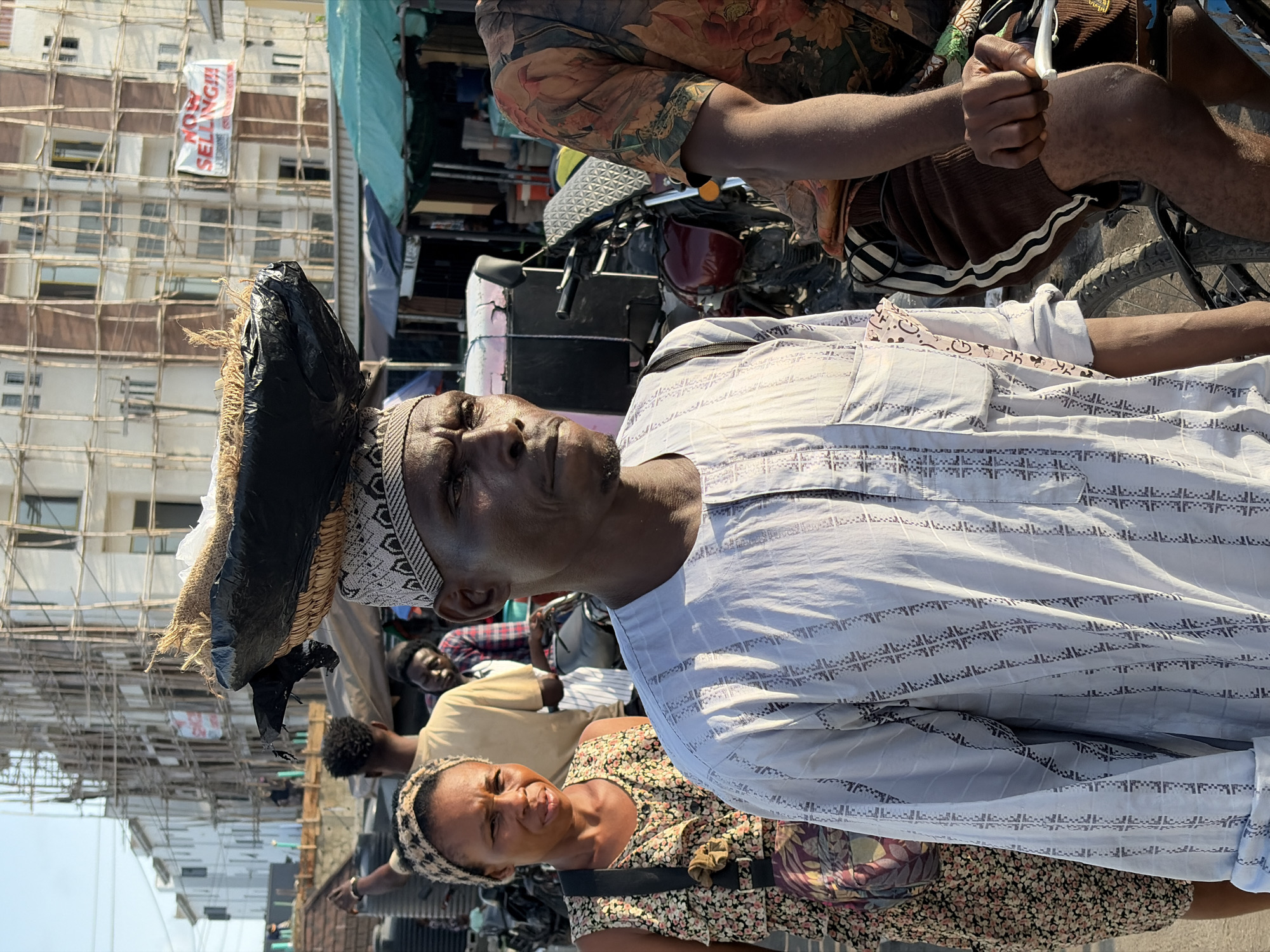 In Igboefon, Nigeria, an eye-level outdoor shot depicts a crowded street. A man in a light blue and white patterned shirt is in the foreground, with a head covering topped by a bundle wrapped in black plastic. Beside him, a woman in a floral dress and headscarf looks towards the camera. Behind them, a man in a light-colored shirt is partially visible. Further back, a motorcycle is parked amidst various objects. A building under construction with bamboo scaffolding dominates the background, displaying a vertical "NOW SELLING!!!" banner. Another man is holding something while sitting on a bicycle. The scene appears to be a bustling marketplace or street scene.
