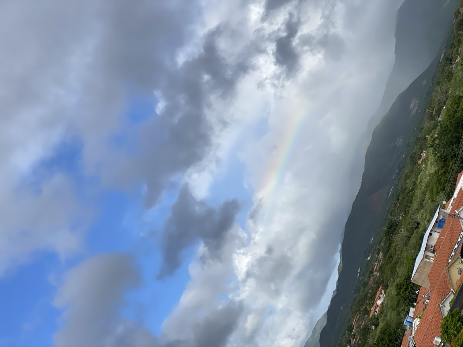 A rainbow arches across a cloudy sky with patches of blue. Gray and white clouds fill most of the frame. A lush, green mountain range slopes down to a residential area with red-tiled roofs and white walls. The buildings are nestled amongst trees. The photograph is rotated counter-clockwise.