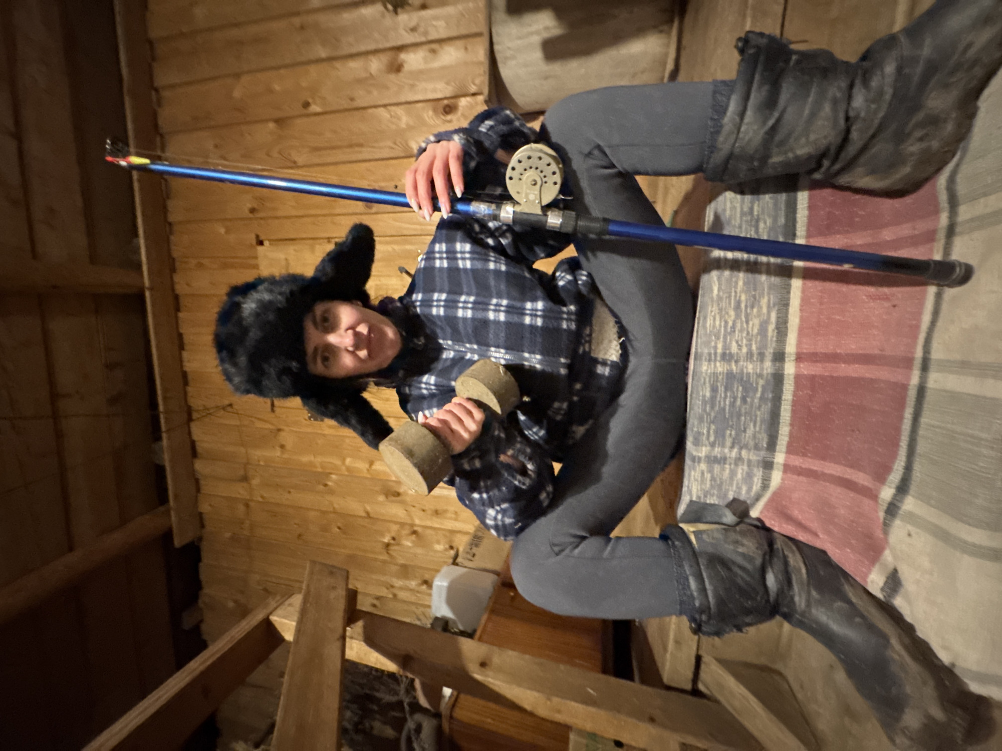 A person is captured indoors in a rustic, wood-paneled room, likely a cabin or shed, with visible wooden plank walls and structural beams. The image is rotated, presenting the scene diagonally.
The individual, appearing to be an adult, is dressed in practical attire suitable for a cold or rural environment. They wear a black fur-style hat with prominent ear flaps, a blue and white plaid flannel shirt, dark grey leggings, and robust, worn black boots. The person looks directly at the camera with a slight smile or smirk, conveying a playful or confident demeanor.
In one hand, they hold a blue fishing rod, complete with a silver fishing reel and small fishing bobbers visible on the line near the tip. In their other hand, they hold a light brown, cylindrical object, which appears to be a homemade weight or dumbbell, possibly crafted from wood or cork.
The background includes a striped blanket or rug, featuring red, grey, and white/cream colors, draped over what looks like a bed or bench. A large, wooden barrel-like object is partially visible in the upper background. The lighting in the room is dim, suggesting either low natural light or reliance on artificial sources. The combination of fishing gear, a makeshift weight, and rustic surroundings hints at a playful or humorous scene, perhaps depicting a blend of outdoor interests and improvised fitness in a rural setting, consistent with a location such as Вассята, Russia. No text is visible in the image.