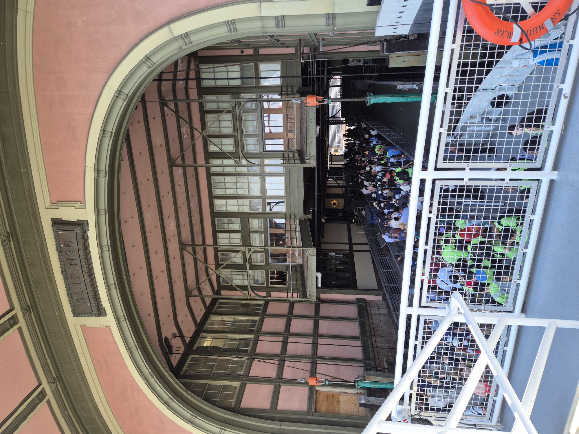 This is an eye-level shot of a ferry terminal entrance, likely at Slip 6 of the Staten Island Ferry terminal in New York City. The scene is viewed from the deck of a ferry, looking towards the land. A large arched opening, painted pink and green with ornate trim, frames the view into the terminal building. Above the arch, a weathered metal sign reads "SLIP N°6".
Inside the terminal, a long line of people are waiting to board a ferry, moving down a ramp. The building's interior is a complex structure of metal beams, girders, and large windows, with a pink ceiling. Numerous cables hang from the ceiling, some attached to orange and green mechanical components.
In the foreground, a white metal railing with a wire mesh barrier separates the viewer from the waiting area. Some of the people in the line are wearing bright green vests, suggesting they might be part of a tour group or organized event. On the right side of the foreground, an orange life preserver is attached to the railing. The overall lighting suggests a bright, sunny day.