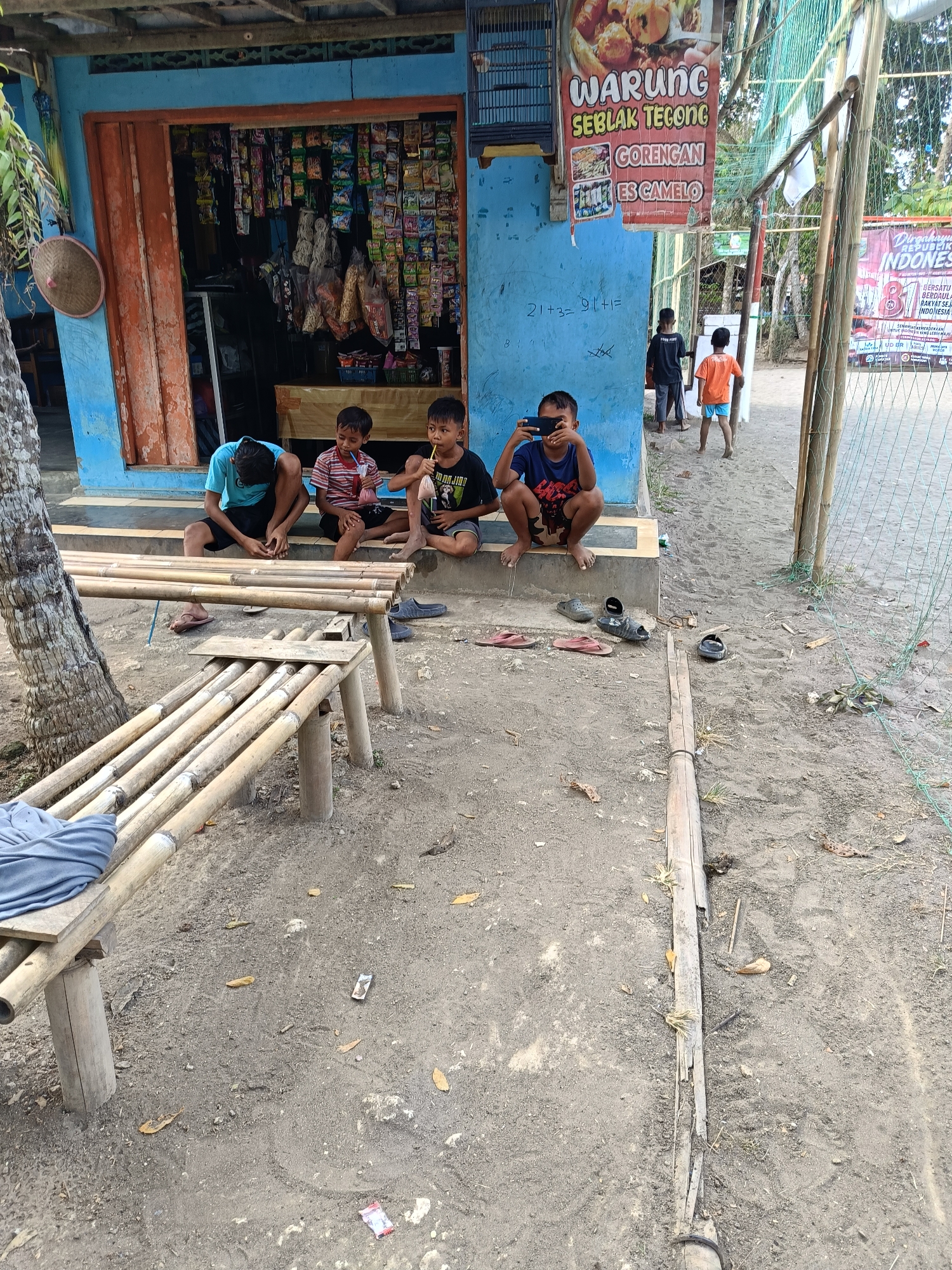 In Buayan, Indonesia, a street-level scene unfolds outside a small shop with a blue facade. Four young boys are the primary subjects, gathered on a concrete step in front of the store. The boy on the far left, wearing a blue t-shirt and black shorts, is crouched down, looking at a black mobile phone in his hands. Next to him, a boy in a striped t-shirt and dark shorts sits, holding a straw to his mouth, possibly drinking from a beverage. To his right, a boy in a black t-shirt and dark shorts is also holding a straw, with a drink in front of him. The fourth boy, partially visible in the background, stands in the doorway of the shop wearing an orange shirt and blue shorts.
The shop itself appears to be a "Warung" selling various goods, as evidenced by the colorful packaging visible through the open doorway. A sign on the wall advertises "WARUNG SEBLAK TEKONG GORENGAN ES CAMELO". The ground in front of the shop is a mixture of sand and dirt, with several pairs of flip-flops scattered around. A long bamboo bench, constructed from several pieces of bamboo, sits in the foreground, with a blue cloth draped over one end. A large tree trunk with visible roots is to the left of the bench, and a woven conical hat hangs on the side of the building. In the background, a net structure, possibly part of a sports court, is visible. The overall atmosphere suggests a casual, outdoor gathering space in a rural or semi-urban Indonesian setting.