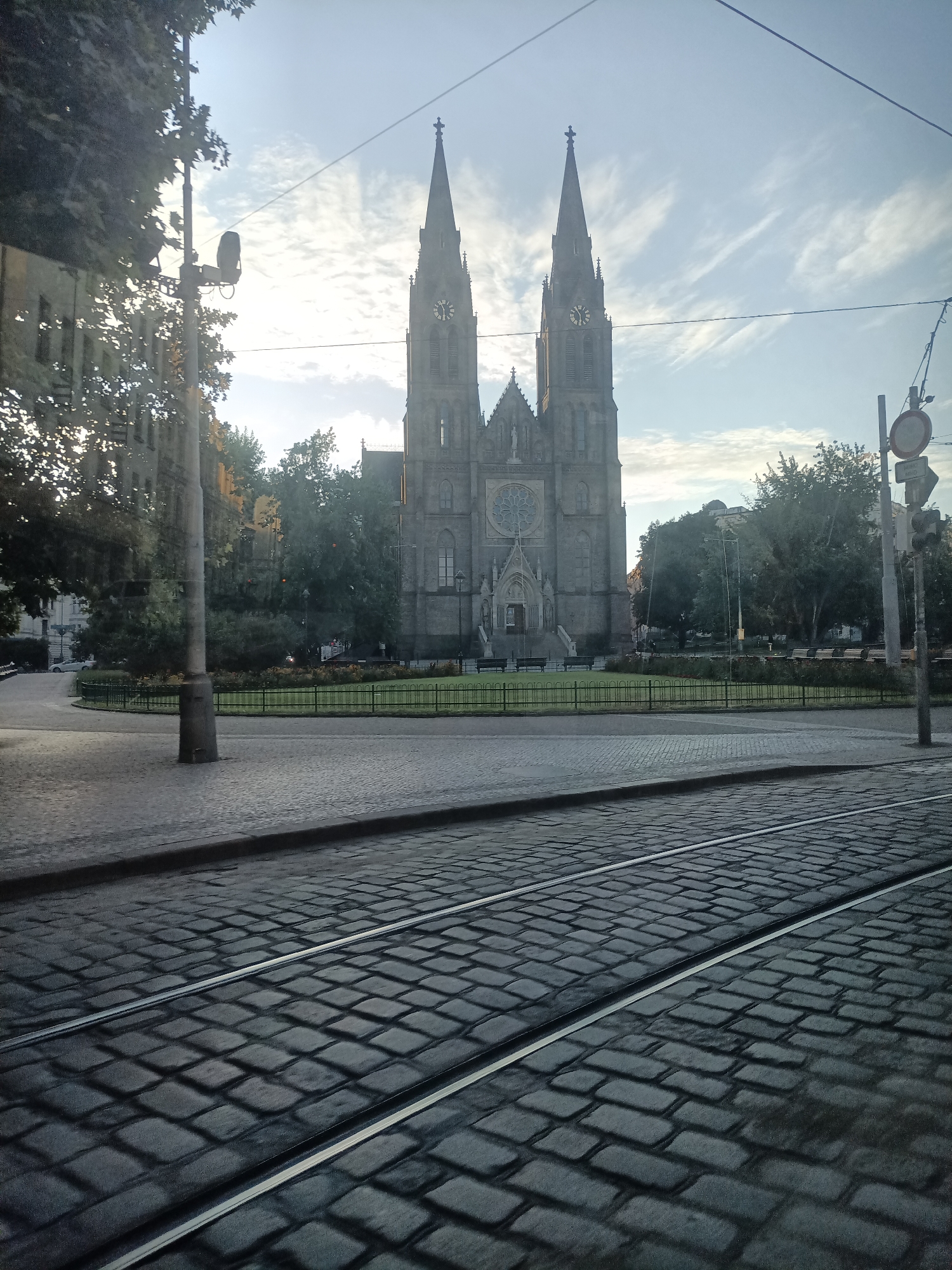 The image shows a view of the Basilica of St. Peter and St. Paul in Vyšehrad, Prague, Czechia. The basilica is a prominent neo-Gothic structure with two tall spires and a large rose window. In the foreground, cobblestone streets with tram tracks are visible. Trees and a neatly manicured lawn with a low fence surround the basilica. The sky is partly cloudy, suggesting it is either early morning or late afternoon. There are no people or animals visible in the scene, and no specific actions or interactions are depicted. The overall impression is one of a quiet, historic urban landscape.