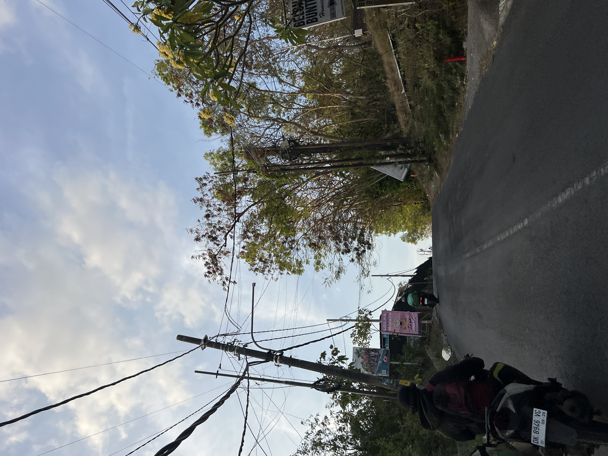 The image depicts a paved road winding uphill in Kuta Selatan, Indonesia, under a partly cloudy sky. In the foreground, a person wearing a black hoodie and a red backpack rides a motorcycle with the license plate "DK 8946 VG". Further up the road, another motorcycle with a rider in a green jacket is visible. The roadside is lined with numerous utility poles and tangled overhead wires. To the right of the road, a sign with the text "BELONGA BUNTU" is partially visible. Dense vegetation, including trees with sparse foliage and some blooming yellow flowers, borders the road. The overall scene suggests a typical day on a slightly rural or less developed road within the region.