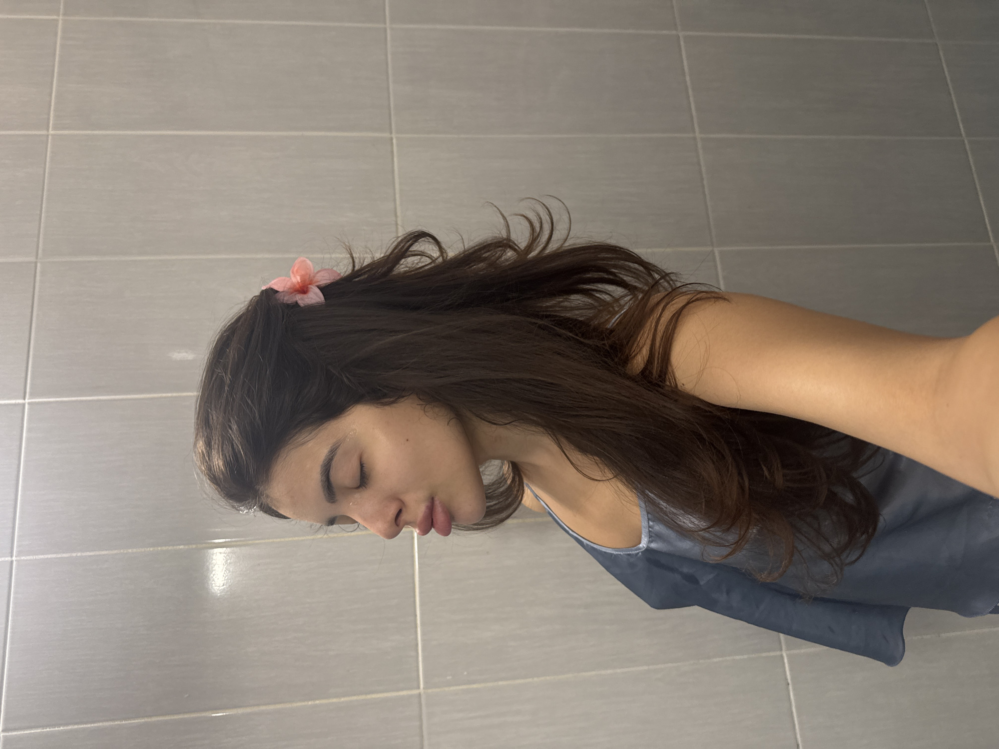 The image shows a medium shot of a young woman with long, dark hair, a pink flower tucked behind her ear, and her eyes closed. She is wearing a blue silk top and appears to be posing with her lips puckered. She is standing against a tiled wall. The location is not specified in the image, and it is not possible to determine if it is in Plovdiv, Bulgaria, or any other city.