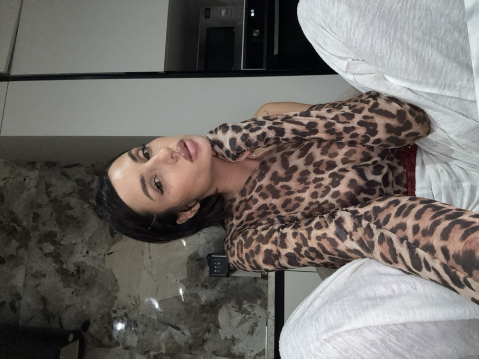 A woman with dark hair and light skin poses indoors. She wears a leopard-print top and light-colored pants, her hand near her neck. The background includes a kitchen countertop with appliances and a wall with a marble-like pattern. The image is slightly rotated.