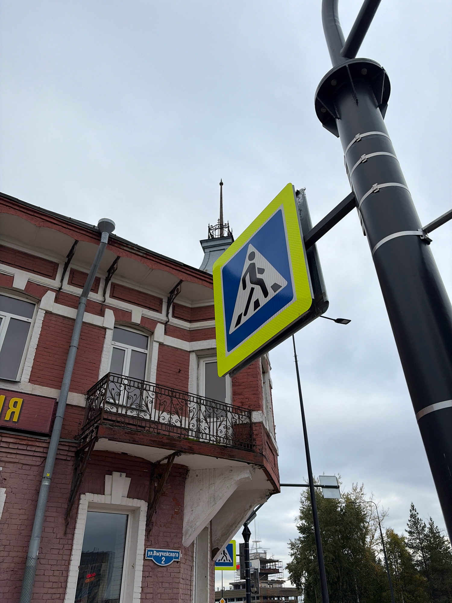 The image captures a street view in Arkhangelsk, Russia, with a pedestrian crossing sign prominently displayed. The sign, a blue square with a white pedestrian silhouette, is mounted on a black metal pole that extends upwards and curves out of frame. The pole itself is secured with several metal bands. To the left of the sign is a portion of a red brick building with white trim around the windows. A metal balcony with an ornate railing juts out from the building. A grey downspout runs vertically along the side of the building. On the building, a blue street sign reads "ул. Вычужского 2" (Vychuzhskogo Street 2). The sky is overcast with grey clouds. In the background, a construction site with scaffolding is visible, along with trees. The overall scene suggests an urban environment on a cloudy day.