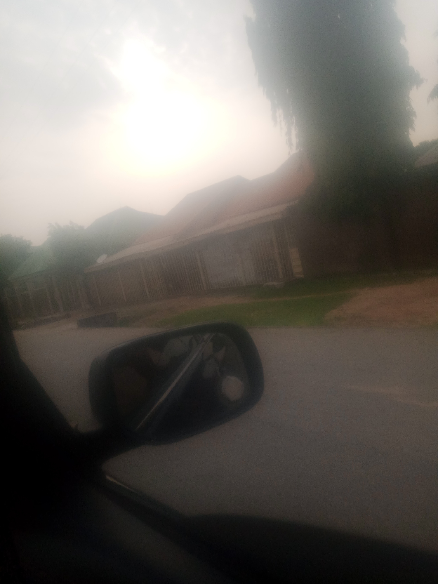 The image depicts a view from inside a vehicle, looking outwards towards a street in Jalingo, Nigeria. A side-view mirror is prominently visible in the foreground on the right, reflecting a portion of the interior of the vehicle and a blurred image of the sky. Beyond the mirror, a road or paved surface stretches into the distance. On the left side of the road, a series of buildings with corrugated iron roofs and walls constructed from vertical wooden slats are visible. Some of these buildings appear to be single-story structures. A patch of green grass borders the buildings and the road. In the background, the sky is overcast and hazy, with a bright, diffused light suggesting daylight, possibly early morning or late afternoon, or a dust-laden atmosphere. A large, dark, out-of-focus tree with drooping branches dominates the right side of the background. The overall impression is of a residential or commercial area in Jalingo, viewed from a moving vehicle under atmospheric conditions that reduce visibility.