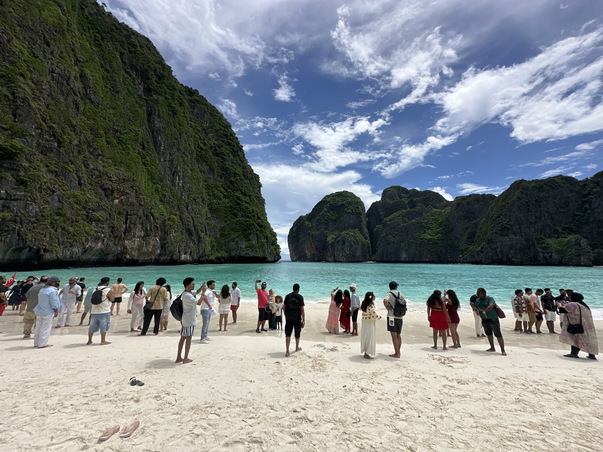 A wide-angle, eye-level shot captures a crowded beach scene in Ao Nang, Thailand, with dramatic, lush green cliffs rising on either side of a turquoise bay. The sand is pale and speckled with footprints and a few pairs of flip-flops. A diverse group of tourists, dressed in casual summer clothing, are scattered across the beach, some posing for photos, others walking, and some conversing. The water is calm and clear, with a distant view of other rocky islands. The sky is a bright blue, adorned with wispy white clouds, suggesting a warm, sunny day.