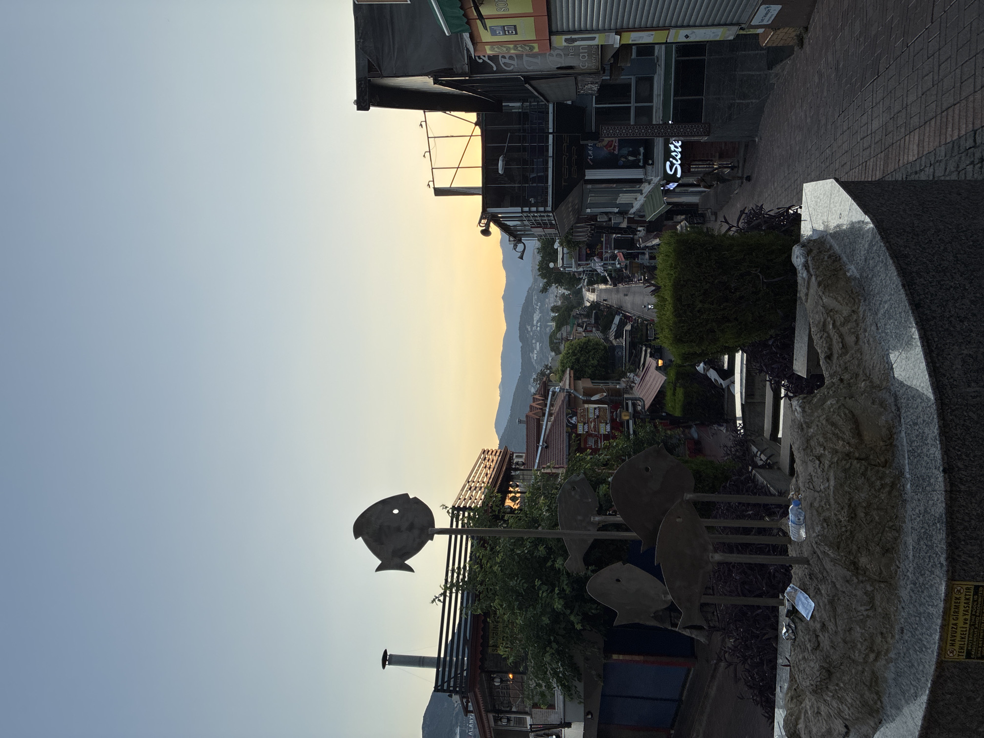 The image depicts a view down a street in Alanya, Turkey, with buildings on either side. The street slopes downwards and is paved with cobblestones. The sky is a clear, pale blue, with a hint of yellow near the horizon, suggesting it is either early morning or late evening. On the left side of the street, there are several buildings with balconies and outdoor seating areas, some adorned with greenery. A prominent metal sculpture of several fish is positioned in the foreground, mounted on a rocky base. The base itself has some litter, including a plastic bottle and a wrapper. A sign at the bottom of the base, written in Turkish, reads "HAVUZA GİRMEK TEHLİKELİ VE YASAKTIR" which translates to "ENTERING THE POOL IS DANGEROUS AND PROHIBITED". Further down the street, the buildings become more commercial, with signs and storefronts visible, including one with neon lettering that appears to spell "Siste". There are a few people visible walking on the street in the distance. On the right side of the street, more buildings are present, with some featuring balconies and awnings. In the background, hills are visible, covered in vegetation. The overall impression is that of a moderately busy street in a coastal town, possibly a tourist area, at a time of day with soft lighting.