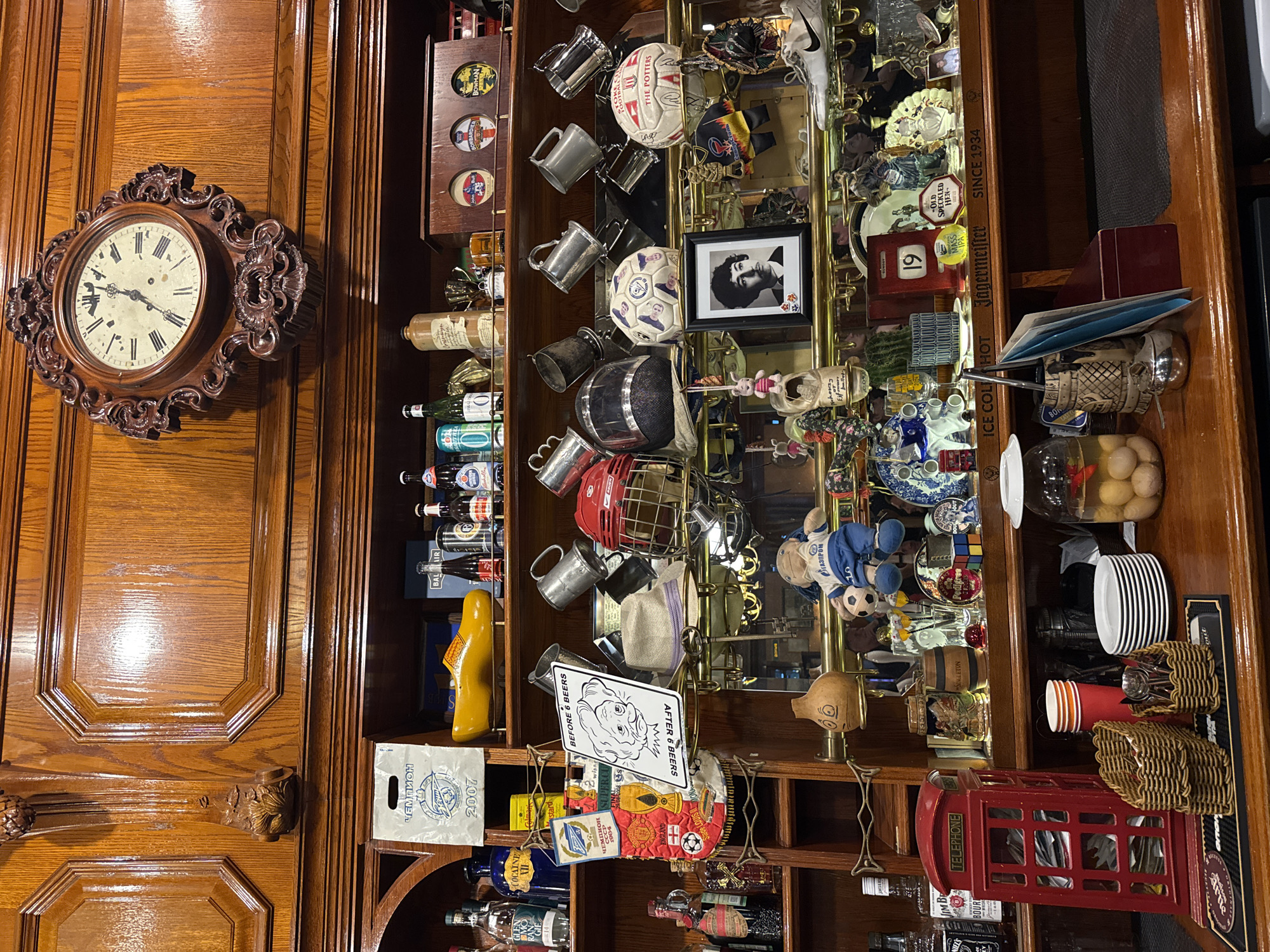 The image depicts a cluttered wooden bar counter and shelves, filled with various objects and memorabilia. On the top shelf, a large ornate clock hangs above a collection of bottles and what appears to be a wooden shoe. The middle shelf displays an assortment of items including mugs, helmets, footballs, a framed photograph, and a plush toy. The lower section of the bar features a red telephone booth replica, various bottles of alcohol, jars with food items, stacks of plates, and wicker baskets. The environment appears to be an indoor establishment, possibly a bar or a pub, with warm wooden paneling and reflective surfaces. There are no people clearly visible in the image. The scene is rich in detail, suggesting a place with a lot of history and collected items. The location is identified as Saint Petersburg, Russia.