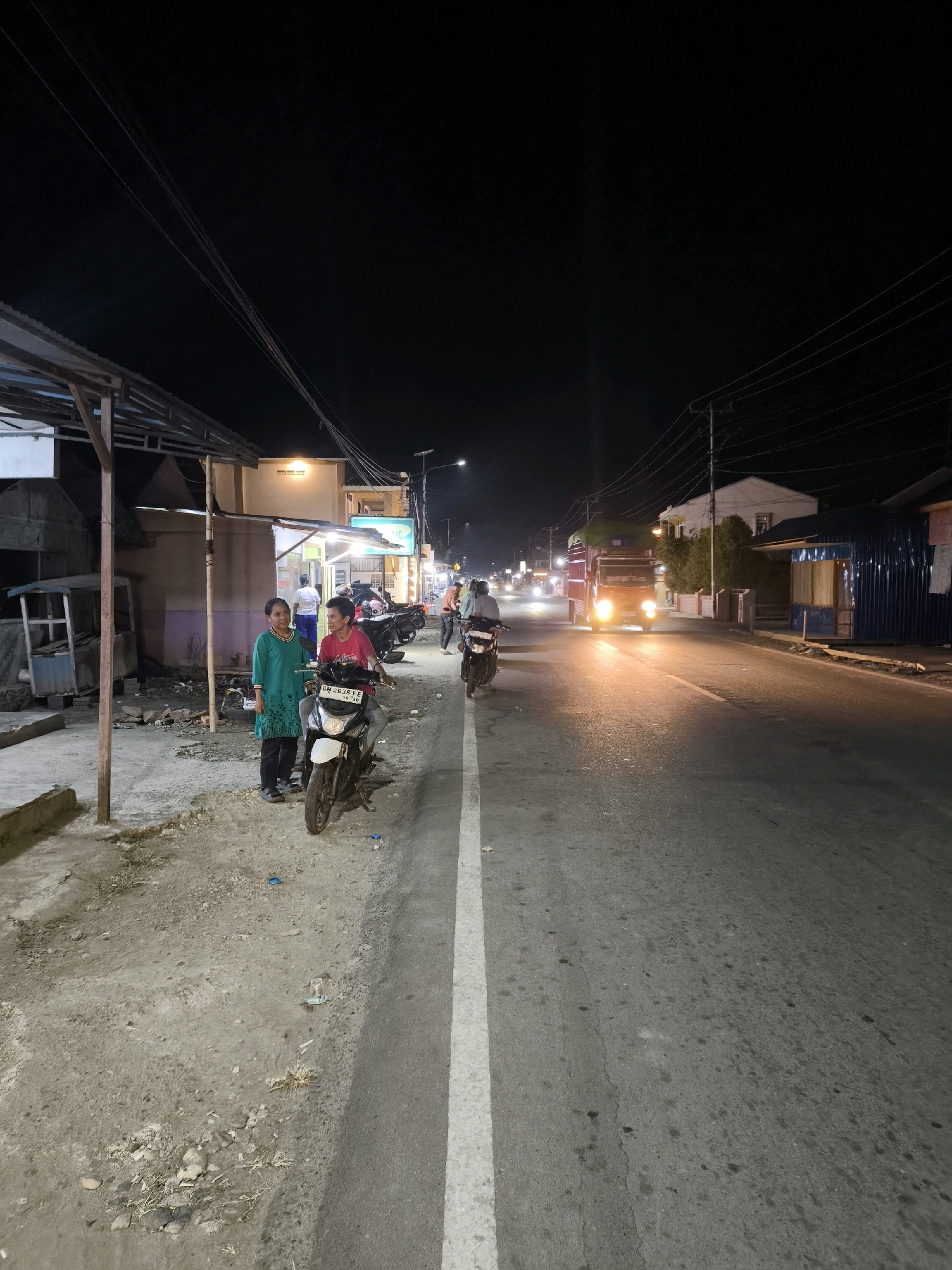 This is a nighttime street scene in Moluo, Indonesia. A paved road with a white dividing line runs through the center of the frame, illuminated by the headlights of passing vehicles and streetlights. On the left side of the road, there is a dirt and gravel shoulder with several structures, including a covered open-air stall with a corrugated metal roof and wooden supports. Two women are standing on the shoulder next to a parked motorcycle. The woman on the left is wearing a green long-sleeved shirt and dark pants. The woman on the right is wearing a red t-shirt and dark pants and is seated on the motorcycle. Behind them, several other motorcycles are parked. Further down the road, a red truck with its headlights on is driving away from the camera. The background shows buildings and trees, with a dark night sky above. The scene appears to be a roadside area with businesses or stalls.
