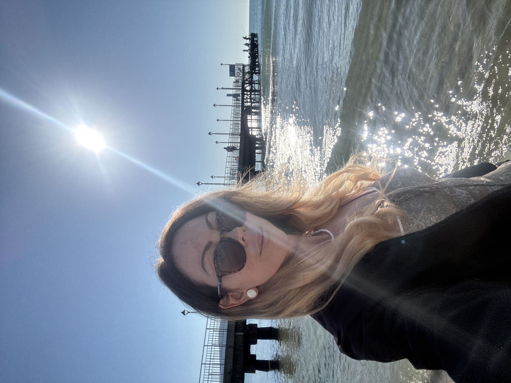 The image shows a woman taking a selfie on a beach in Nesebar, Bulgaria, with a pier in the background. The sun is bright and high in the sky, casting a strong glare on the water and pier. The woman is wearing sunglasses, a black jacket over a patterned top, and a white necklace. She has blonde-streaked hair and is wearing a large, light-colored earring. The pier, a long wooden structure supported by pilings, extends into the sea. Several people can be seen on the pier, some of whom appear to be fishing. The water is calm with gentle waves, and the sun's reflection creates a shimmering effect on the surface. The sky is clear and blue.