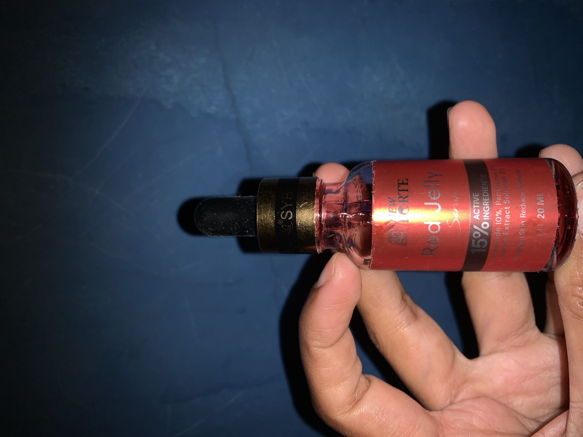 A hand holds a small, red glass bottle with a dropper. The bottle has a gold band around its neck, with some text visible on it. The bottle contains a red liquid, and a label on the bottle reads "NEW FORTE Red Jelly Serum" with "15% ACTIVE INGREDIENTS" below it. The background is a dark blue, textured surface. No specific location in Karangkemiri, Indonesia is identifiable.
