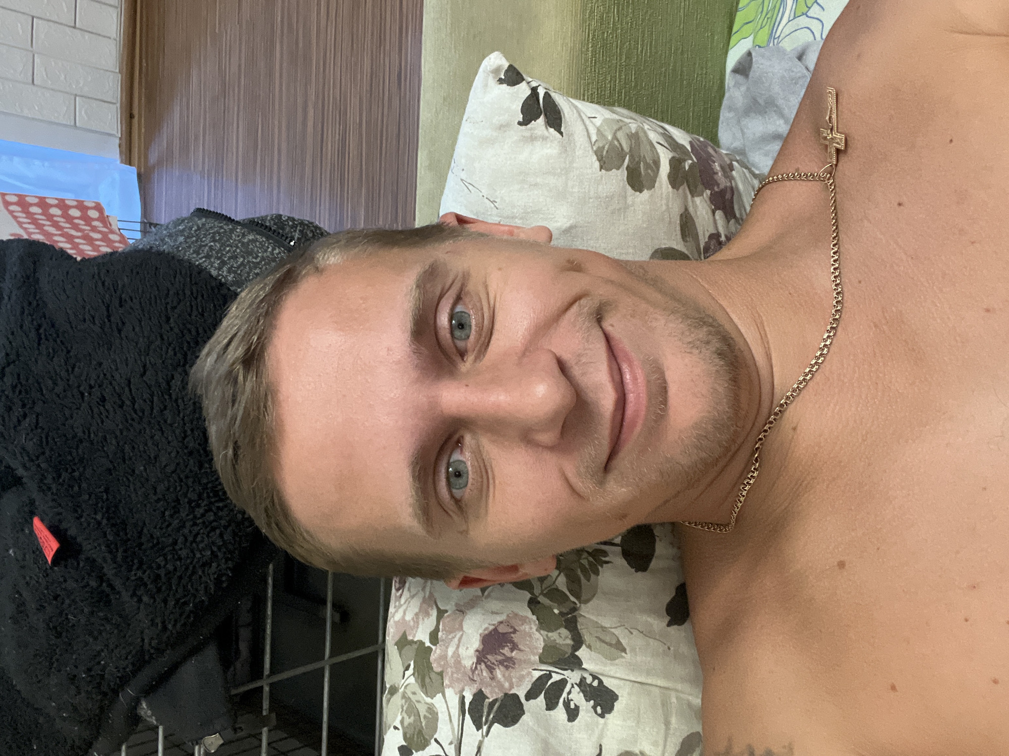 The image is a selfie taken from a low angle by a light-skinned man with blond hair and blue eyes. He is lying down, with his head resting on a floral-patterned pillow. He is wearing a gold chain with a cross pendant. The background consists of a wooden floor, a wall with a light blue and red polka-dot pattern, and a black fuzzy garment. The scene appears to be indoors, likely in a bedroom. No specific location in Minsk, Belarus is identifiable from the image.