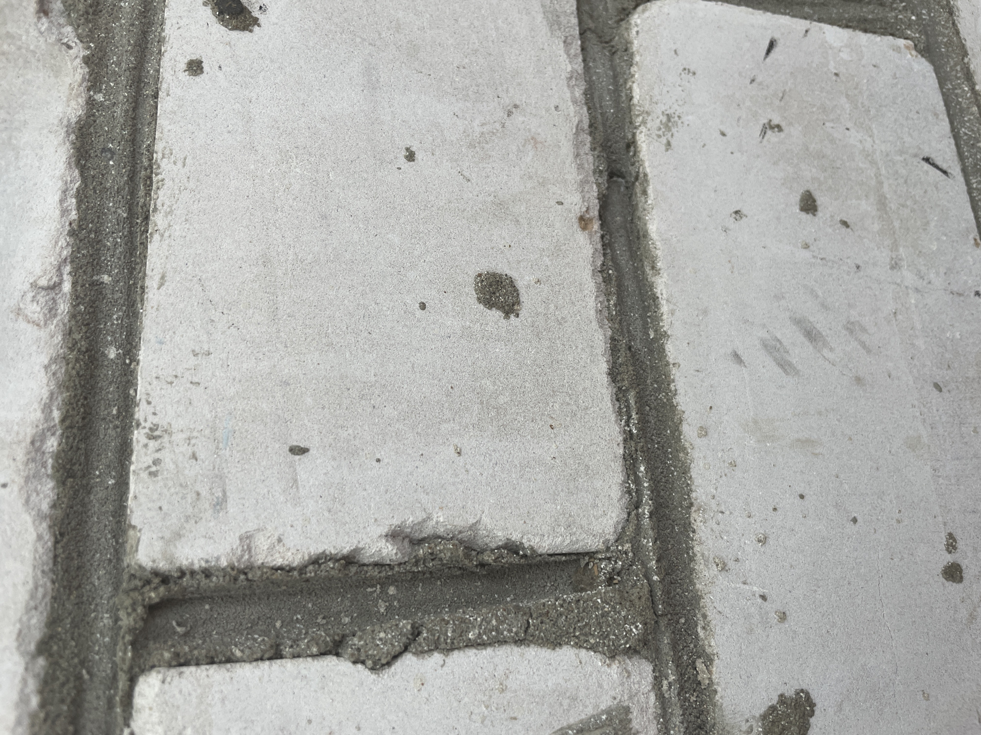The image shows a close-up view of a weathered white brick wall with dark gray mortar. The bricks have an uneven, slightly rough texture and are stained with scattered dark spots and smudges. The mortar is visibly rough and uneven in places, with some chipped or broken areas revealing aggregate. The lighting suggests an outdoor environment, with subtle shadows indicating the direction of light. The scene is consistent with the exterior of a building, potentially an older structure, in an urban or semi-urban setting. Without further context, a precise location in Kotlas, Russia, cannot be determined from the image alone.